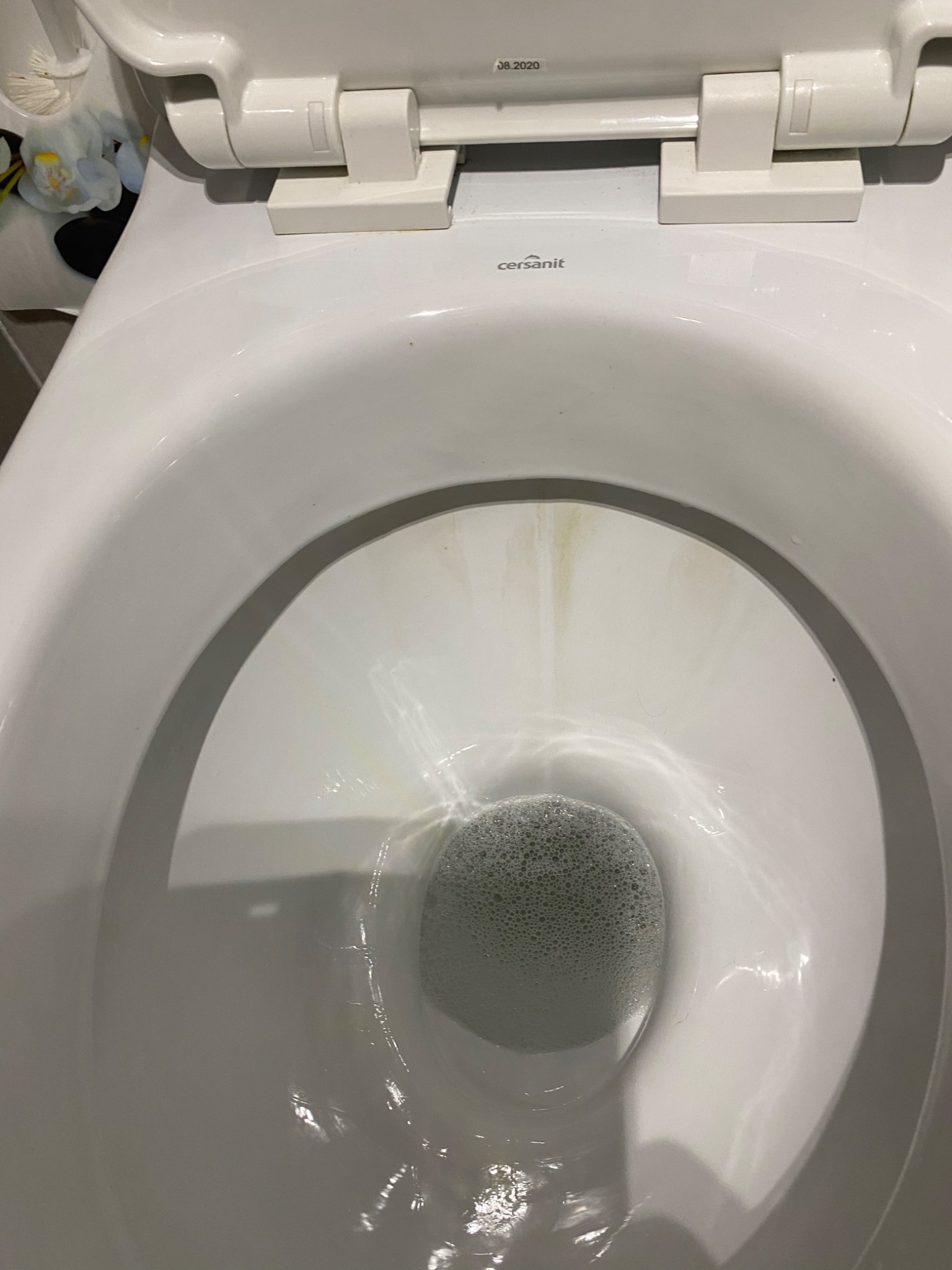 Widok z góry na białą muszlę klozetową z widocznymi smugami zabrudzeń i pianą w wodzie, z otwartą deską sedesową marki Cersanit.