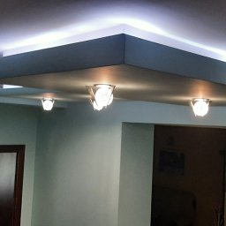 Dariusz Stasiak firma uslugowo-remontowa - Wielopoziomowy sufit podwieszany z oświetleniem LED i trzema ozdobnymi lampami punktowymi, widoczne fragmenty ścian w odcieniach zieleni i drzwi z ciemnego drewna z przeszkleniami.