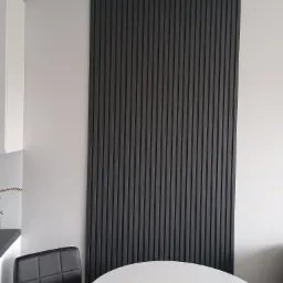 Nowoczesna aranżacja ściany z ciemnymi lamelami w pionowym układzie, widoczna w tle biała ściana i fragment białego stołu. Minimalistyczny design wnętrza.