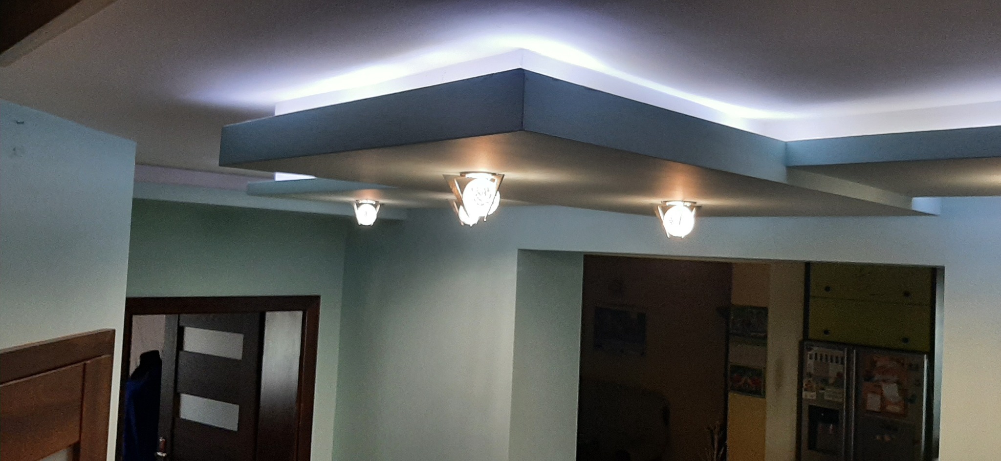 Wielopoziomowy sufit podwieszany z oświetleniem LED i trzema ozdobnymi lampami punktowymi, widoczne fragmenty ścian w odcieniach zieleni i drzwi z ciemnego drewna z przeszkleniami.