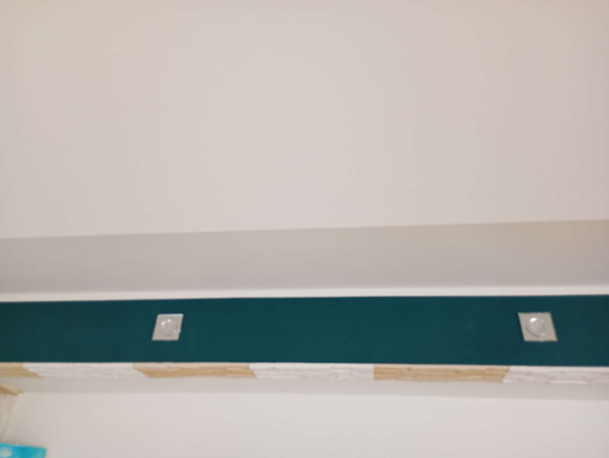 Fragment ściany z pomalowanym na ciemnozielono pasem pod sufitem, widoczne dwa kwadratowe punkty oświetleniowe i biała ściana poniżej z dekoracyjnym pasem płytek.