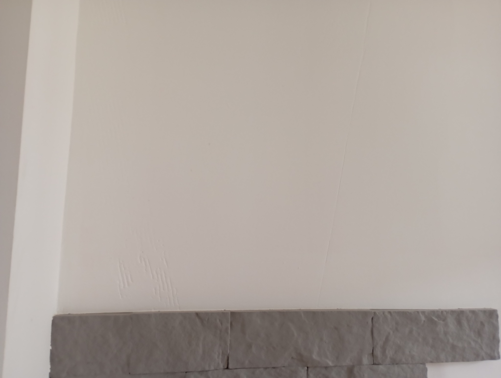 Fragment ściany z nierówną, świeżo pomalowaną powierzchnią, widoczne smugi pędzla i dolny pas z dekoracyjnych, szarych płytek imitujących kamień.