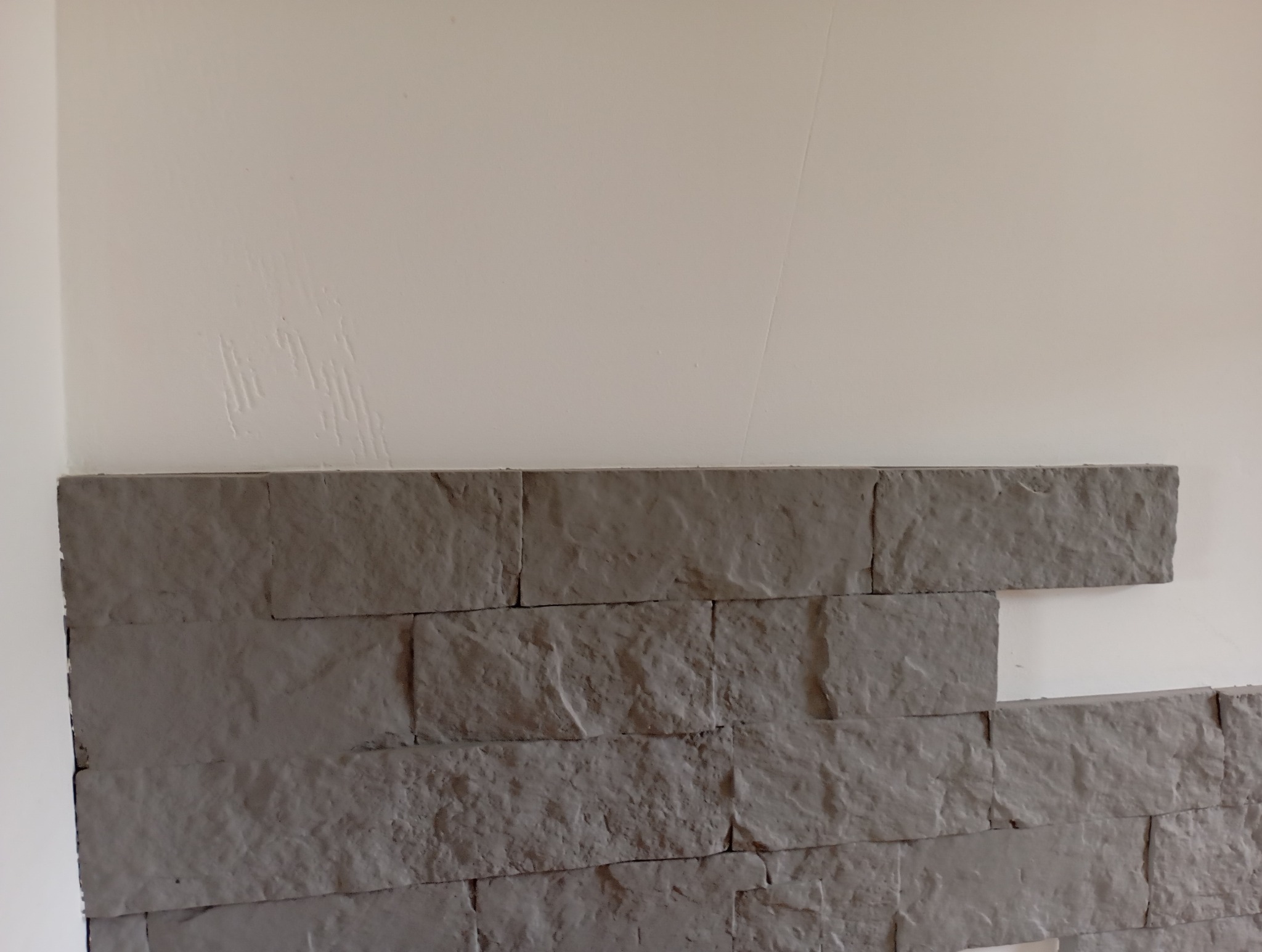 Fragment ściany z szarymi panelami imitującymi kamień, ułożonymi do połowy wysokości, z widocznymi śladami po szpachlowaniu na górnej, białej części ściany.