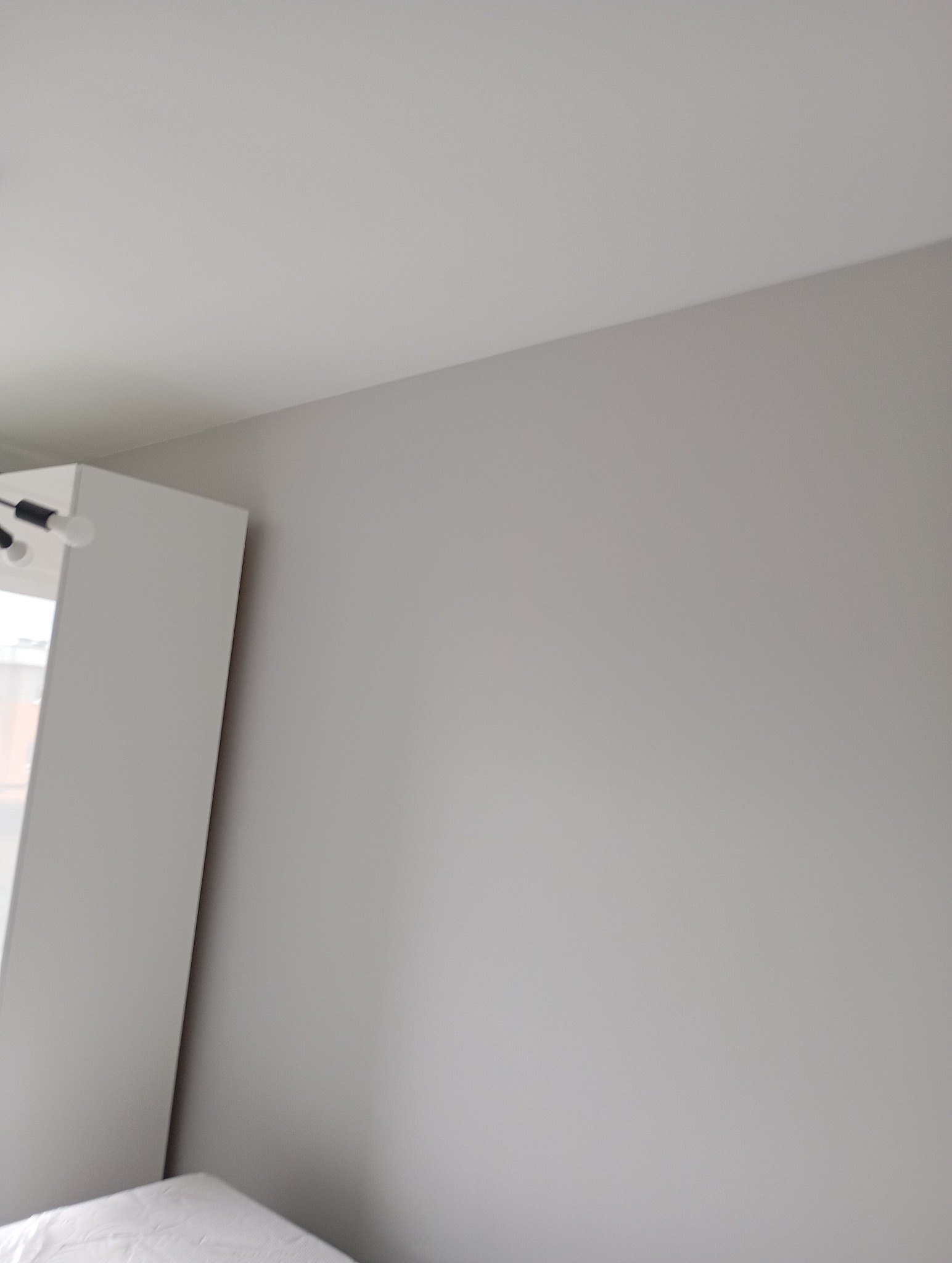 Gładka, szara ściana po malowaniu, widoczny fragment białej szafki i lampy sufitowej. Minimalistyczne wnętrze w neutralnych barwach.