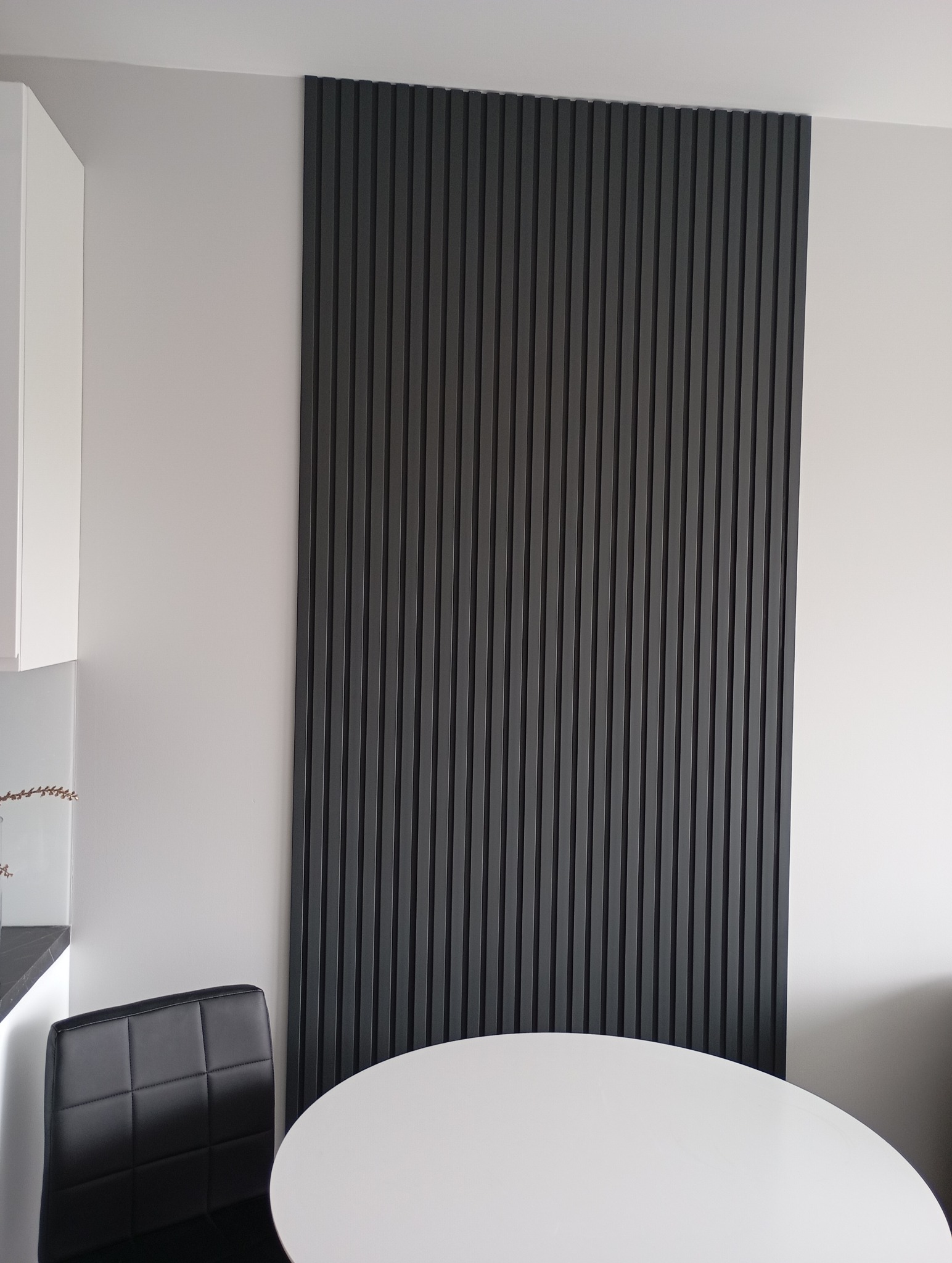 Nowoczesna aranżacja ściany z ciemnymi lamelami w pionowym układzie, widoczna w tle biała ściana i fragment białego stołu. Minimalistyczny design wnętrza.