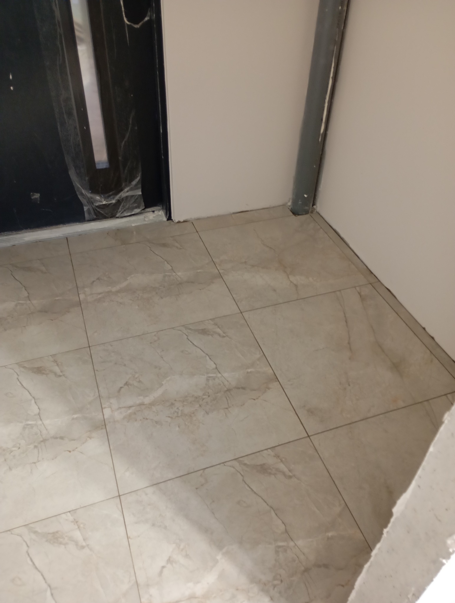 Ułożone, beżowe płytki podłogowe z marmurkowym wzorem, widoczne przy ścianie i futrynie drzwi z folią ochronną. Szara rura przy ścianie.