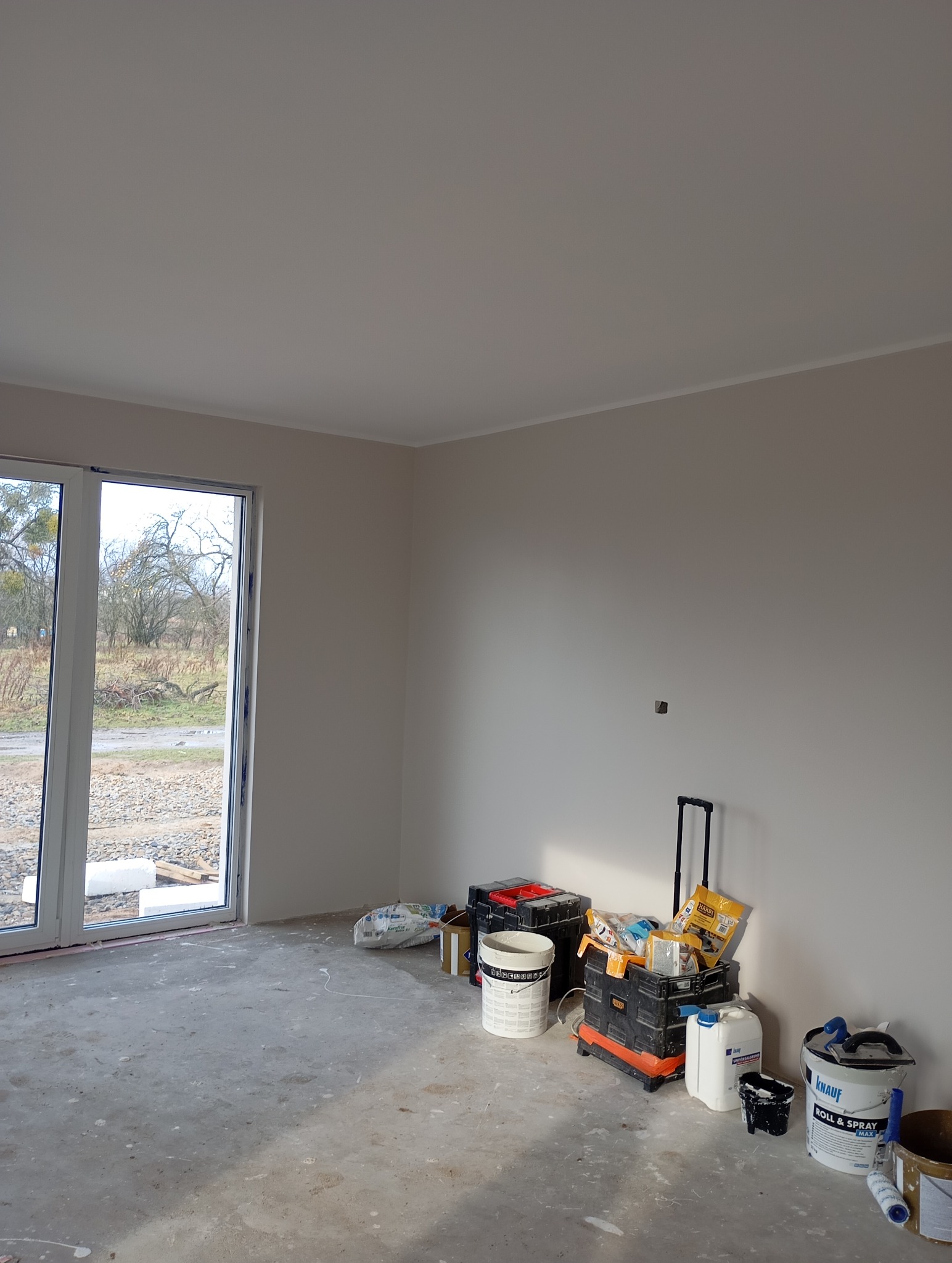 Wnętrze po malowaniu ścian na jasny odcień szarości, widoczne narzędzia malarskie i worki z materiałami budowlanymi. Duże okno z widokiem na zewnątrz.