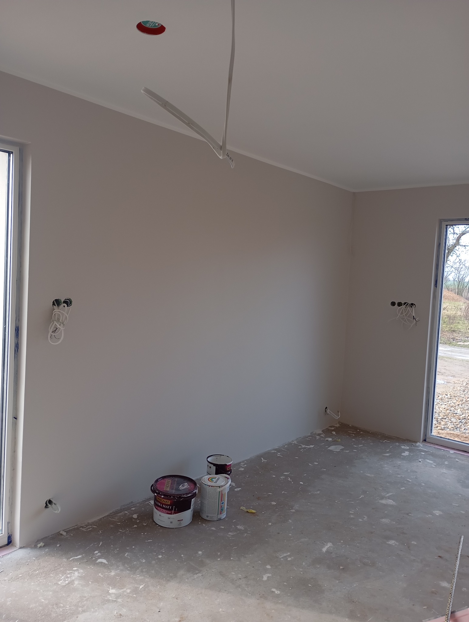 Jasne wnętrze po malowaniu ścian na beżowo, widoczne puszki z farbą na podłodze, przygotowane gniazdka elektryczne i surowy beton. Dwa okna wpuszczają naturalne światło.