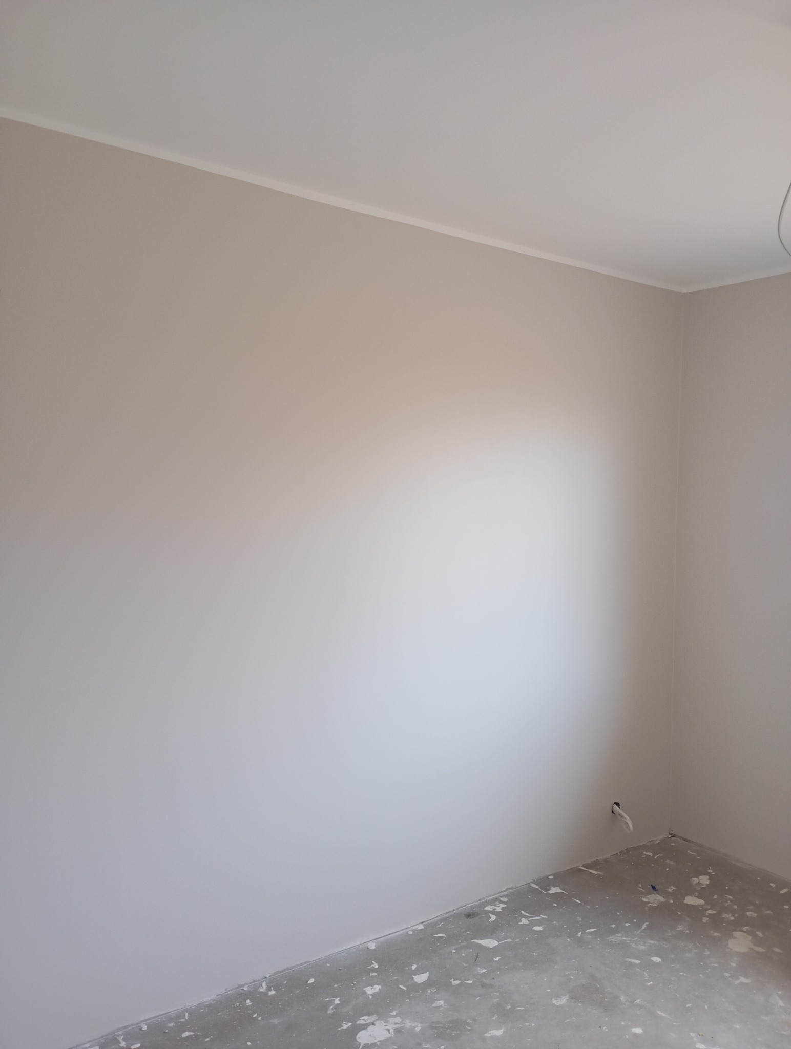 Pomalowane na beżowo ściany w rogu pokoju z białą listwą sufitową. Podłoga w trakcie przygotowania do dalszych prac wykończeniowych. Widoczne gniazdko elektryczne.