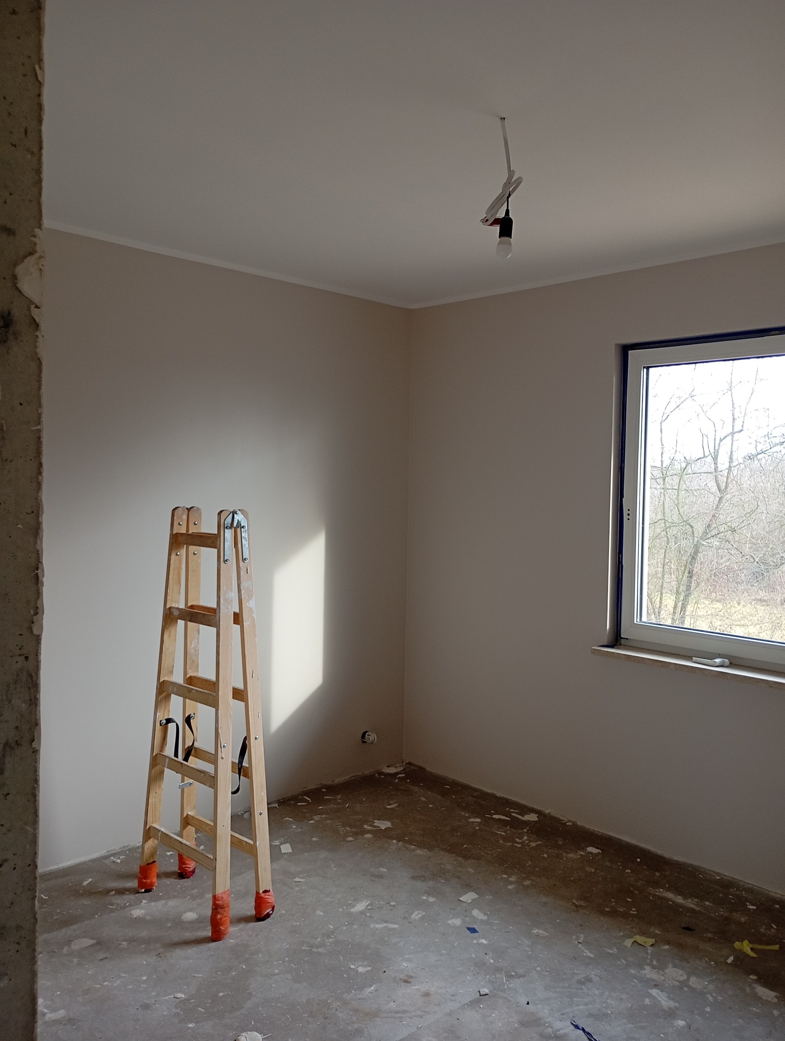 Jasne wnętrze po malowaniu ścian na beżowo, z drewnianą drabiną z pomarańczowymi zabezpieczeniami na nogach i oknem z widokiem na drzewa.