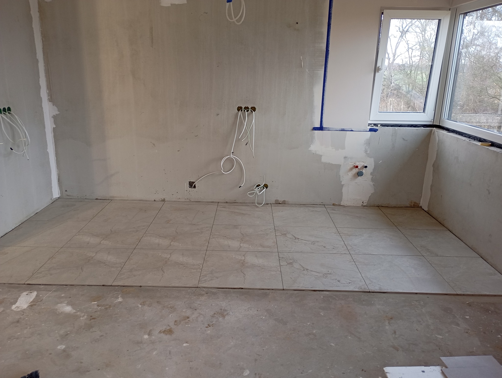 Połowa podłogi wyłożona beżowymi płytkami w trakcie remontu. Na ścianach widoczne kable elektryczne i ślady po gładzi. Okno z widokiem na drzewa w tle.