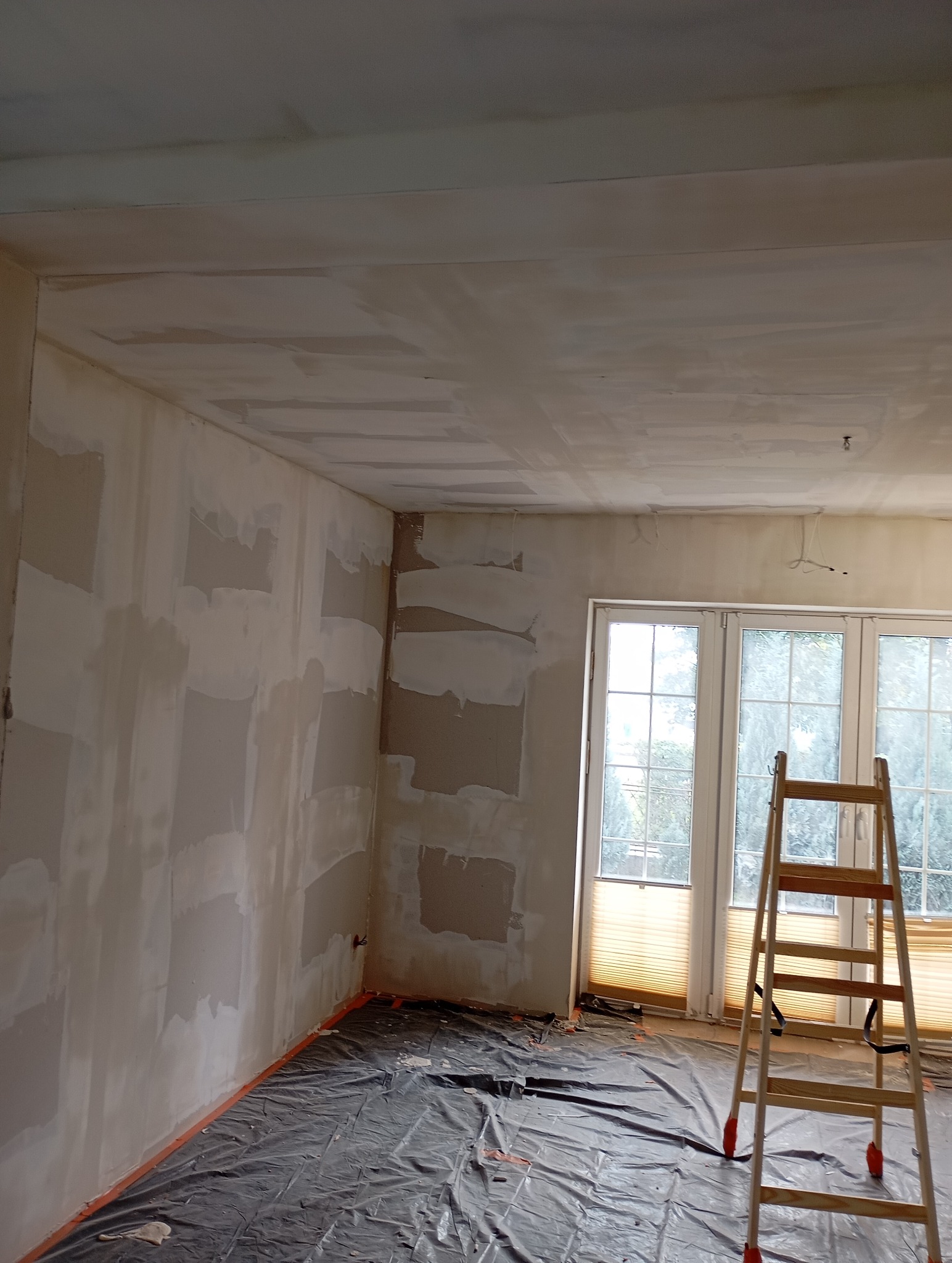 Pokój w trakcie szpachlowania ścian i sufitu, zabezpieczona podłoga folią, drabina oparta o okno. Widoczne niedoskonałości i nierówności na powierzchniach.