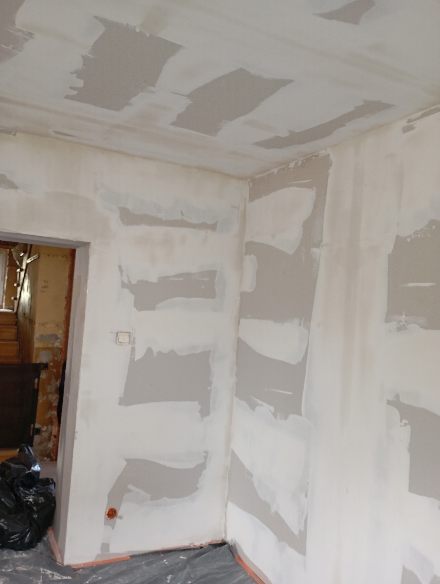 Ściana i sufit w trakcie szpachlowania, widoczne fragmenty nałożonej gładzi. Otwór drzwiowy z widocznym wnętrzem pomieszczenia w tle. Podłoga zabezpieczona folią.