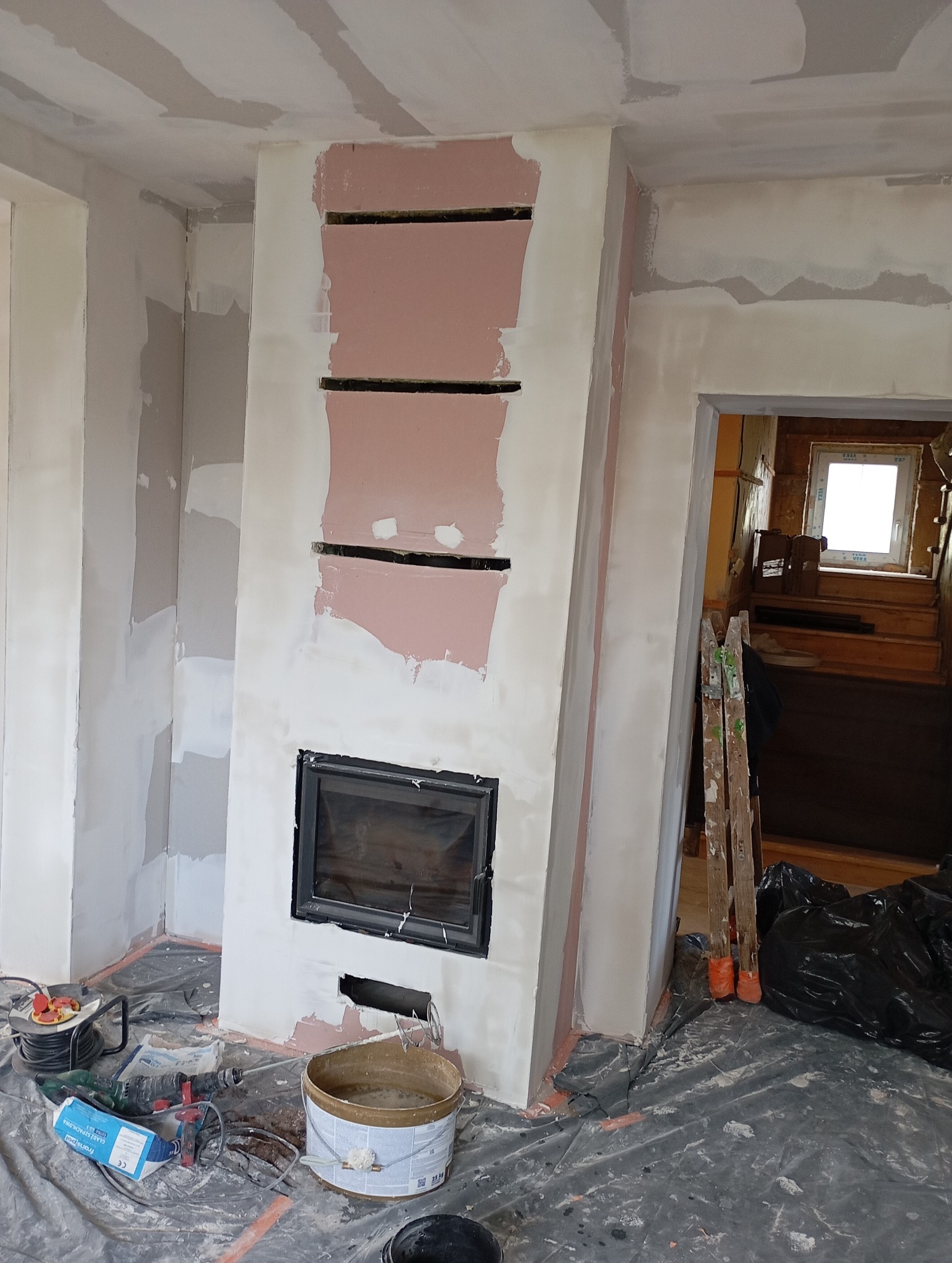 Wnętrze w trakcie prac wykończeniowych: obudowa kominka z płyt karton-gips, szpachlowane ściany, narzędzia i wiadro z zaprawą na podłodze zabezpieczonej folią.