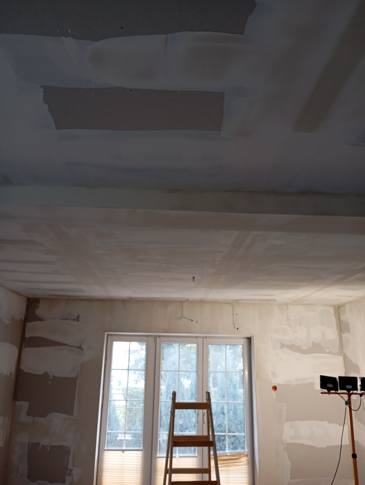 Pokój w trakcie szpachlowania: ściany i sufit pokryte gładzią, drabina oparta o okno, lampa budowlana. Widoczne niedoskonałości i nierówności przed malowaniem.