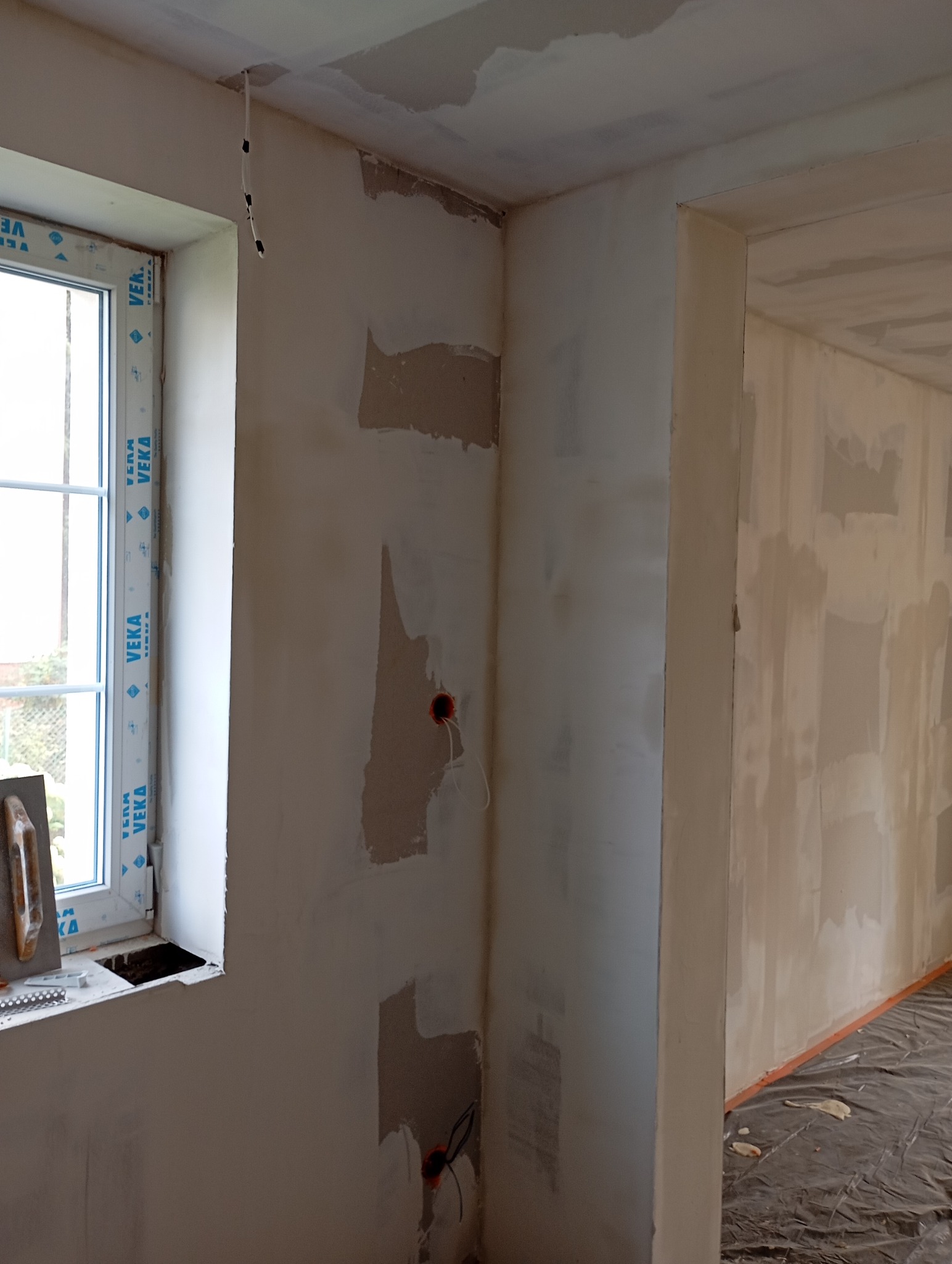 Ściana po szpachlowaniu, widoczne gniazdka elektryczne, okno z narzędziami. Przygotowanie do malowania, nierówności na ścianie, surowy stan wykończenia wnętrza.