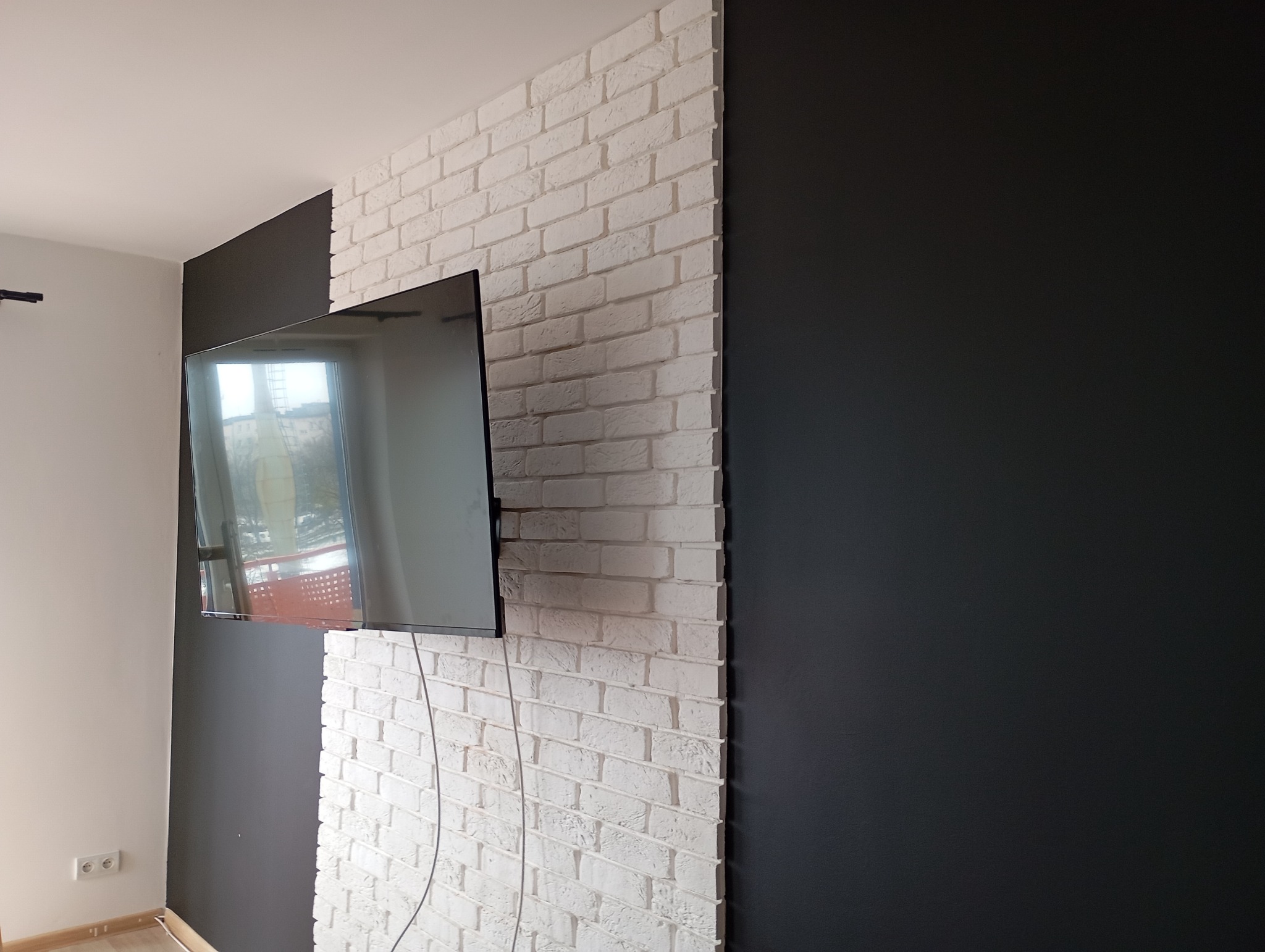 Ściana z białej cegły dekoracyjnej, obok czarna ściana, na ścianie telewizor. Widoczne gniazdka elektryczne. Nowoczesne wykończenie wnętrza.