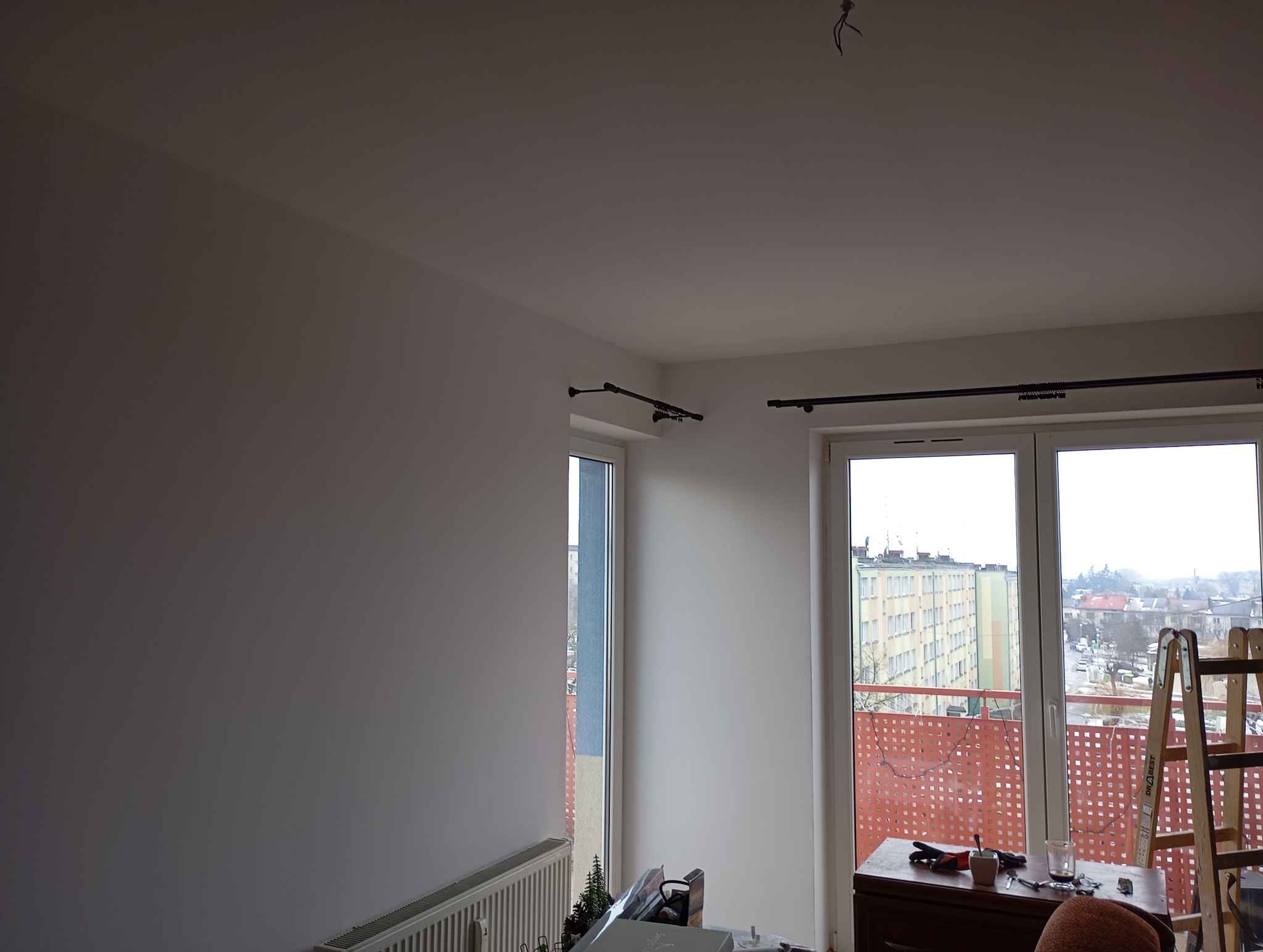 Świeżo pomalowane na biało wnętrze z zamontowanymi karniszami, widok na balkon i drabinę. Minimalistyczny wystrój, jasne ściany, przygotowanie do zawieszenia zasłon.