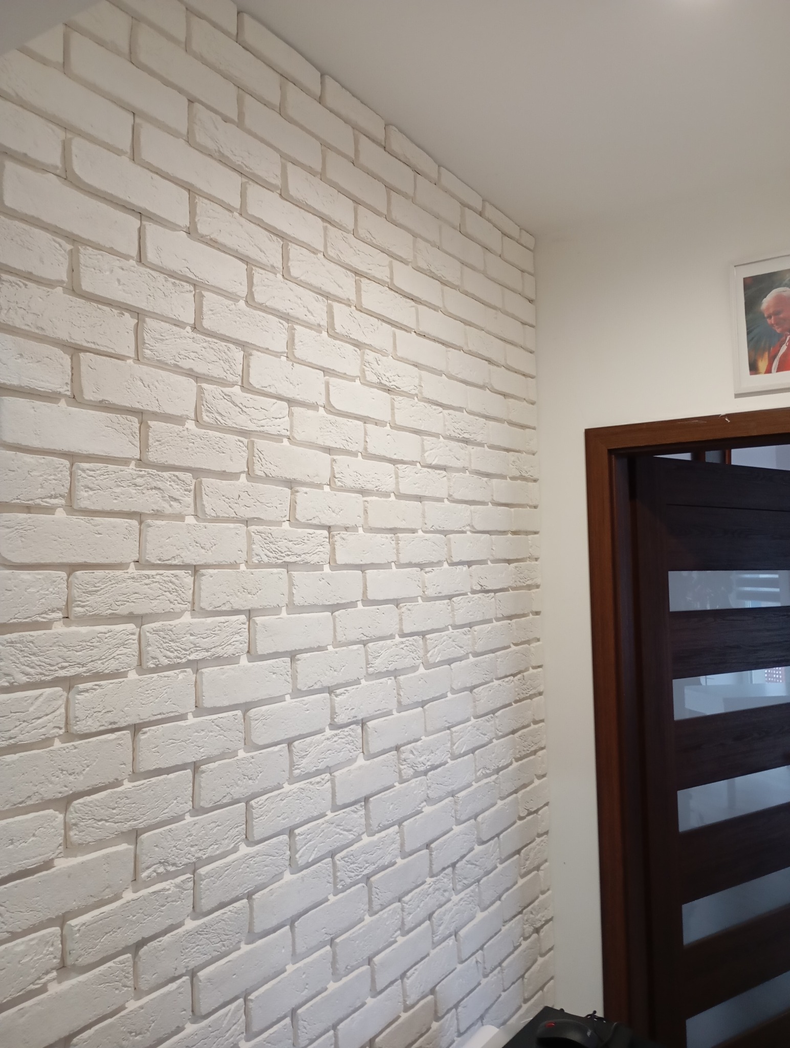 Ściana wykończona białą cegłą dekoracyjną, widok z boku, z fragmentem ciemnych drzwi po prawej stronie. Na ścianie wisi obraz w białej ramie.