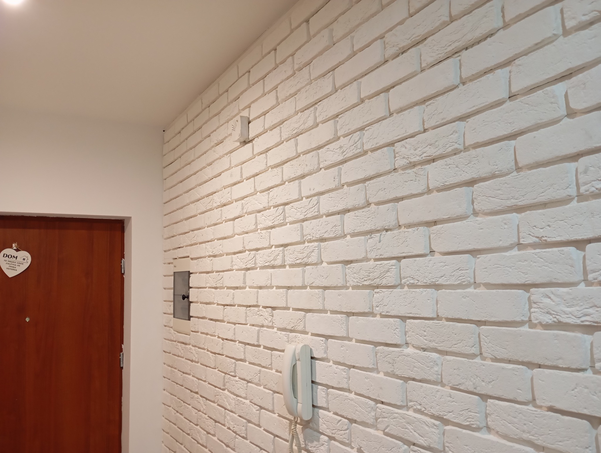 Biała ściana z cegły dekoracyjnej w korytarzu, z widocznym panelem elektrycznym i telefonem stacjonarnym. Obok brązowe drzwi z ozdobą w kształcie serca.