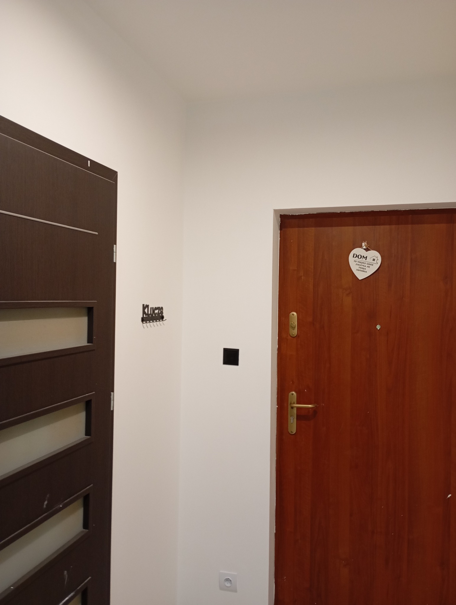 Korytarz po remoncie: białe ściany, ciemne drzwi z szybami, wieszak na klucze i drewniane drzwi z ozdobą w kształcie serca. Minimalistyczny design.