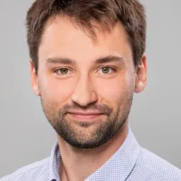 Portret młodego mężczyzny z brązowymi włosami i zarostem, ubranego w błękitną koszulę w drobny wzór, na neutralnym szarym tle.