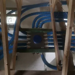 Widok z dołu na rozdzielacz z licznymi niebieskimi rurami karbowanymi, zamontowany w drewnianej konstrukcji stropu poddasza.