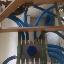 Rozdzielacz instalacji wentylacyjnej z niebieskimi karbowanymi rurami, widoczny w trakcie montażu w przestrzeni poddasza z drewnianą konstrukcją więźby dachowej.