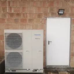 Zewnętrzna jednostka pompy ciepła Panasonic Inverter zamontowana przy ścianie z czerwonej cegły kratówki obok białych drzwi z czarną klamką i czarnym reflektorem nad nimi.