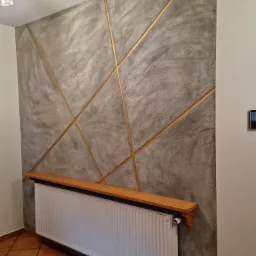 Ściana z szarą dekoracyjną fakturą i złotymi liniami, biały grzejnik z drewnianą półką, terakota na podłodze.