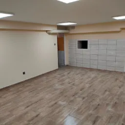 Puste pomieszczenie po remoncie z ułożoną podłogą z płytek imitujących drewno, częściowo wykafelkowaną ścianą z otworem wentylacyjnym, pomalowanymi ścianami na beżowo i białymi drzwiami.