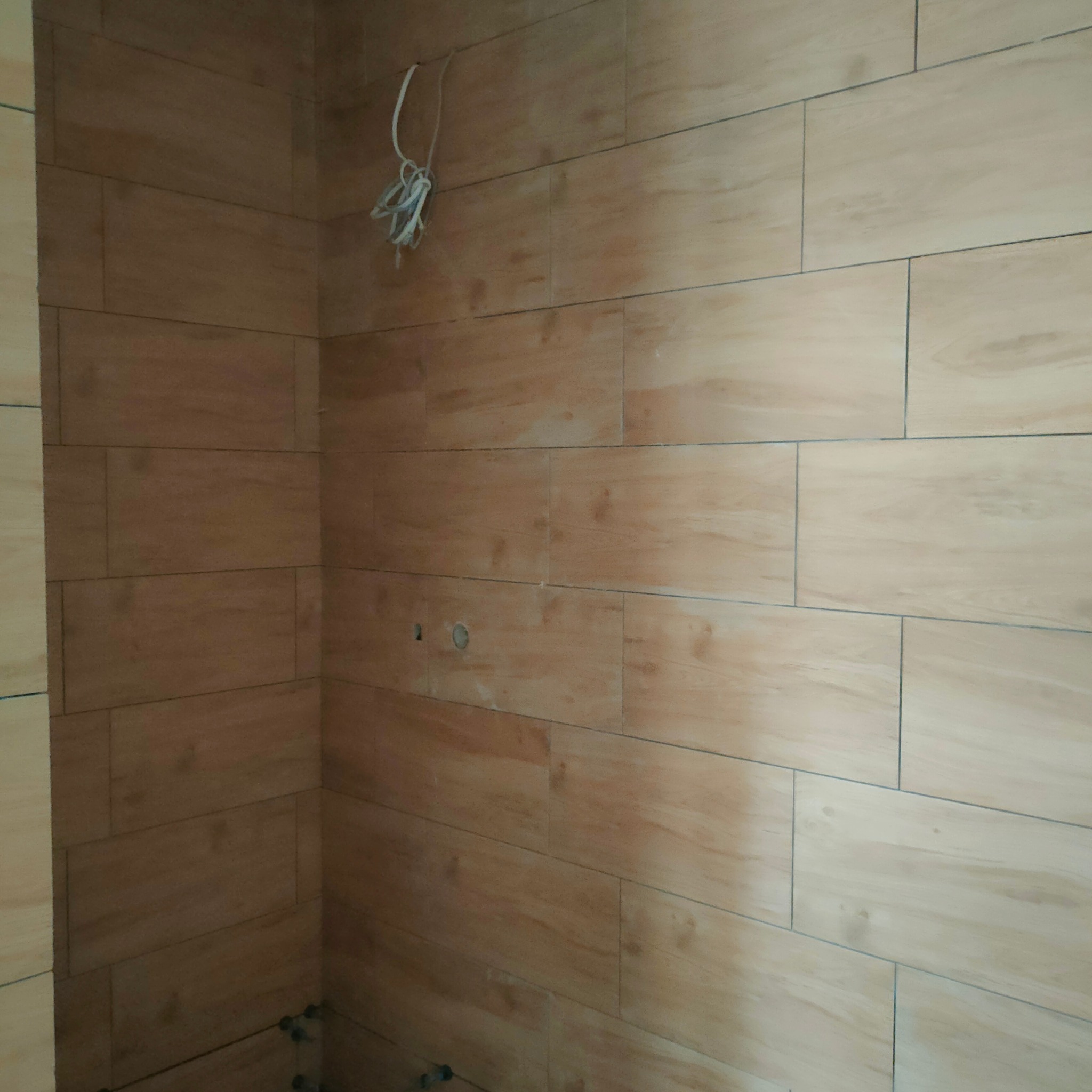 Ściana w trakcie wykańczania z beżowymi, drewnopodobnymi płytkami ceramicznymi ułożonymi poziomo; widoczne wystające przewody elektryczne i otwory na instalacje.