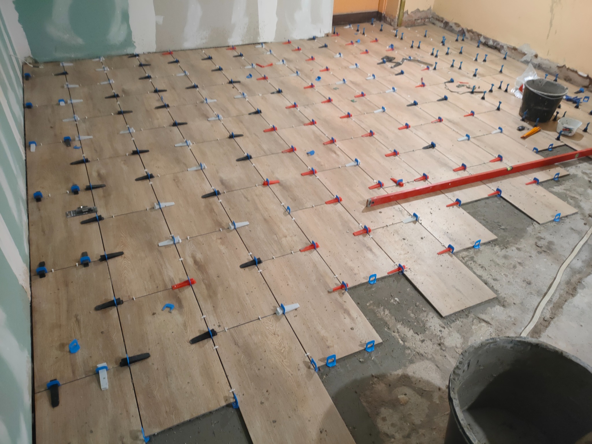 Układanie płytek podłogowych imitujących drewno z użyciem systemu poziomowania, widoczne narzędzia i wiaderka z zaprawą.