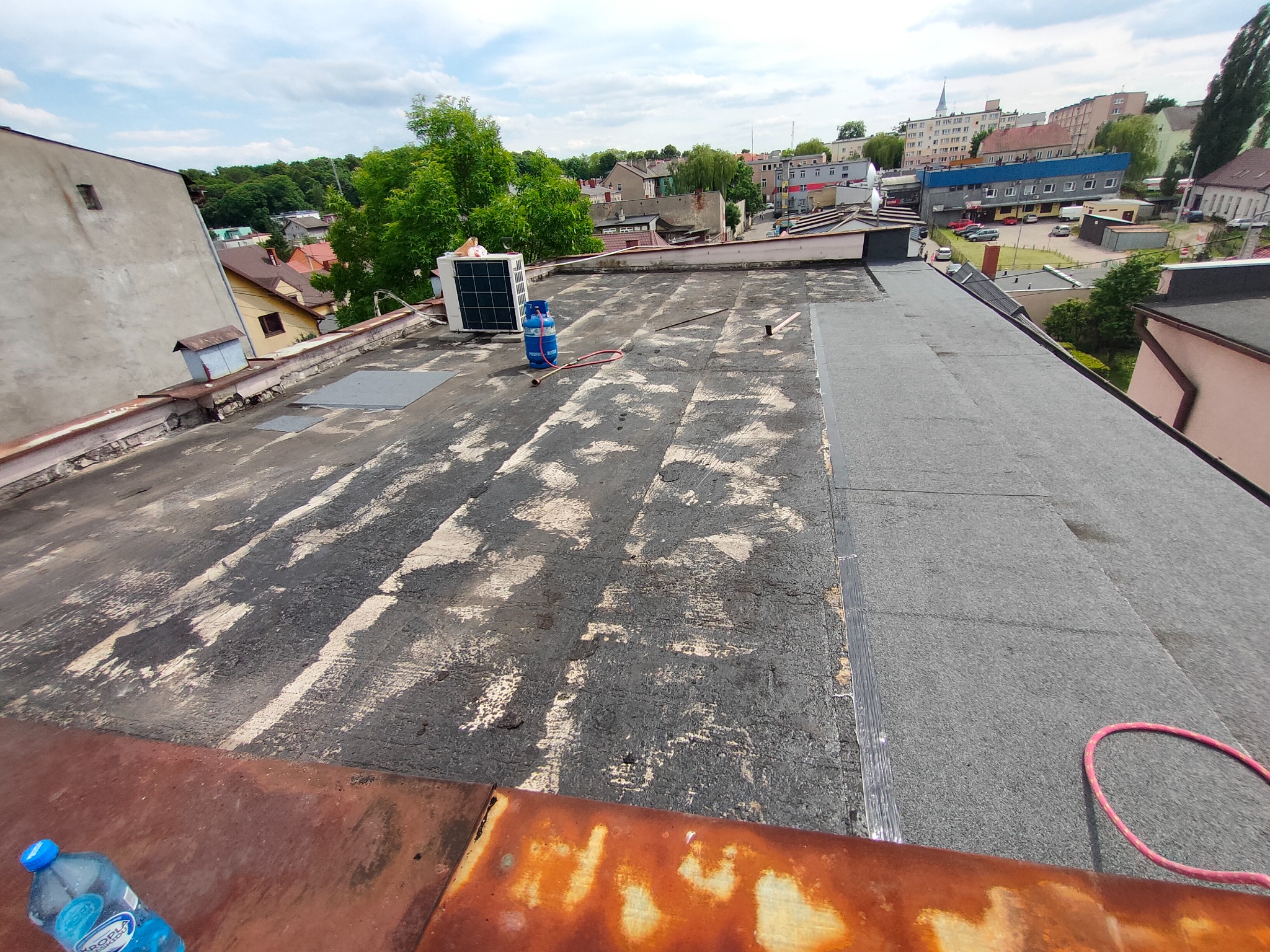 Nierówna powierzchnia dachu pokryta papą z widocznym sprzętem do klimatyzacji, butlą gazową i akcesoriami, w tle zabudowa miejska.