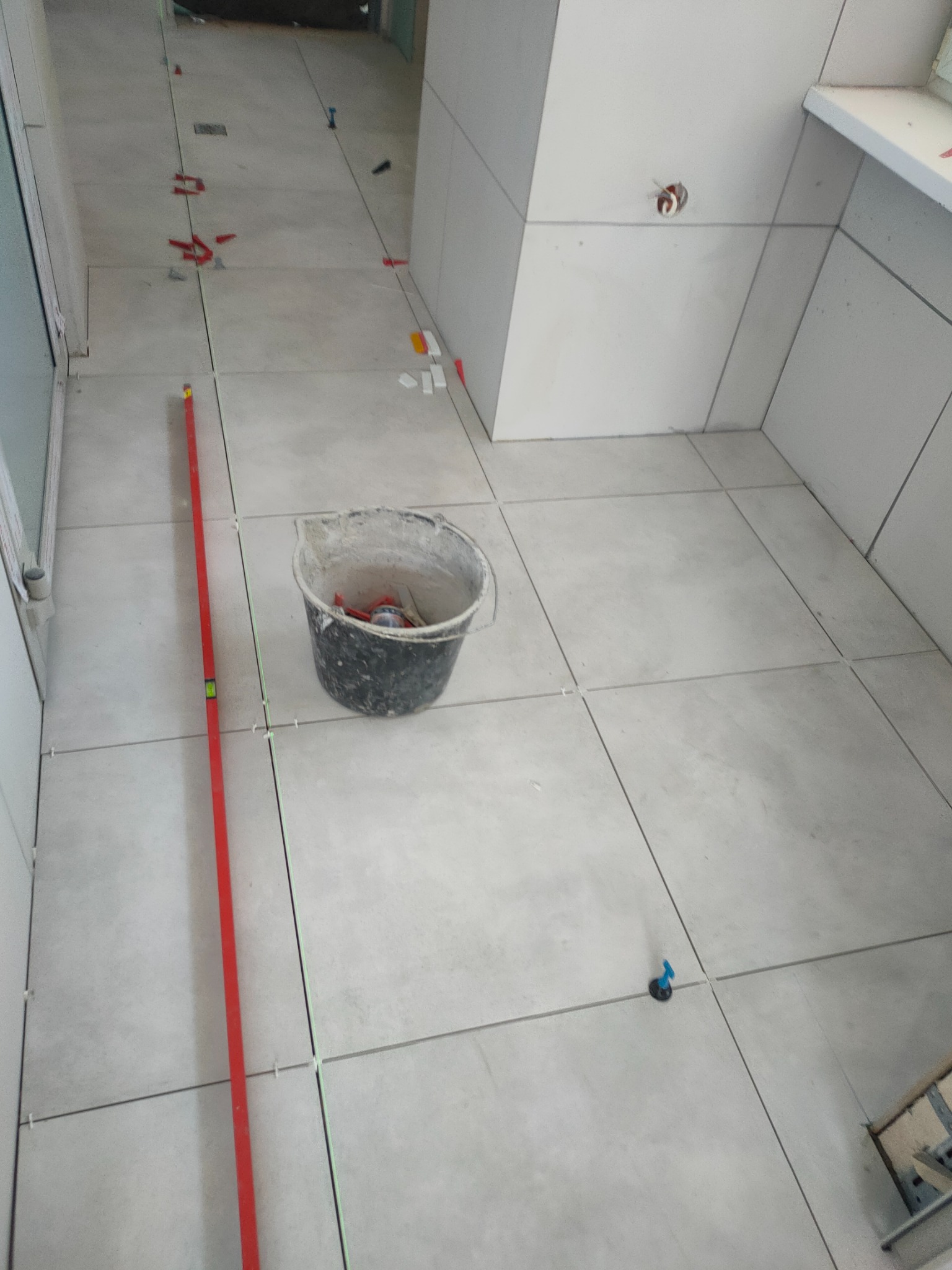 Wnętrze w trakcie kładzenia jasnych, kwadratowych płytek podłogowych i ściennych, widoczny poziomica, wiadro z narzędziami, krzyżyki dystansowe i system poziomowania płytek.