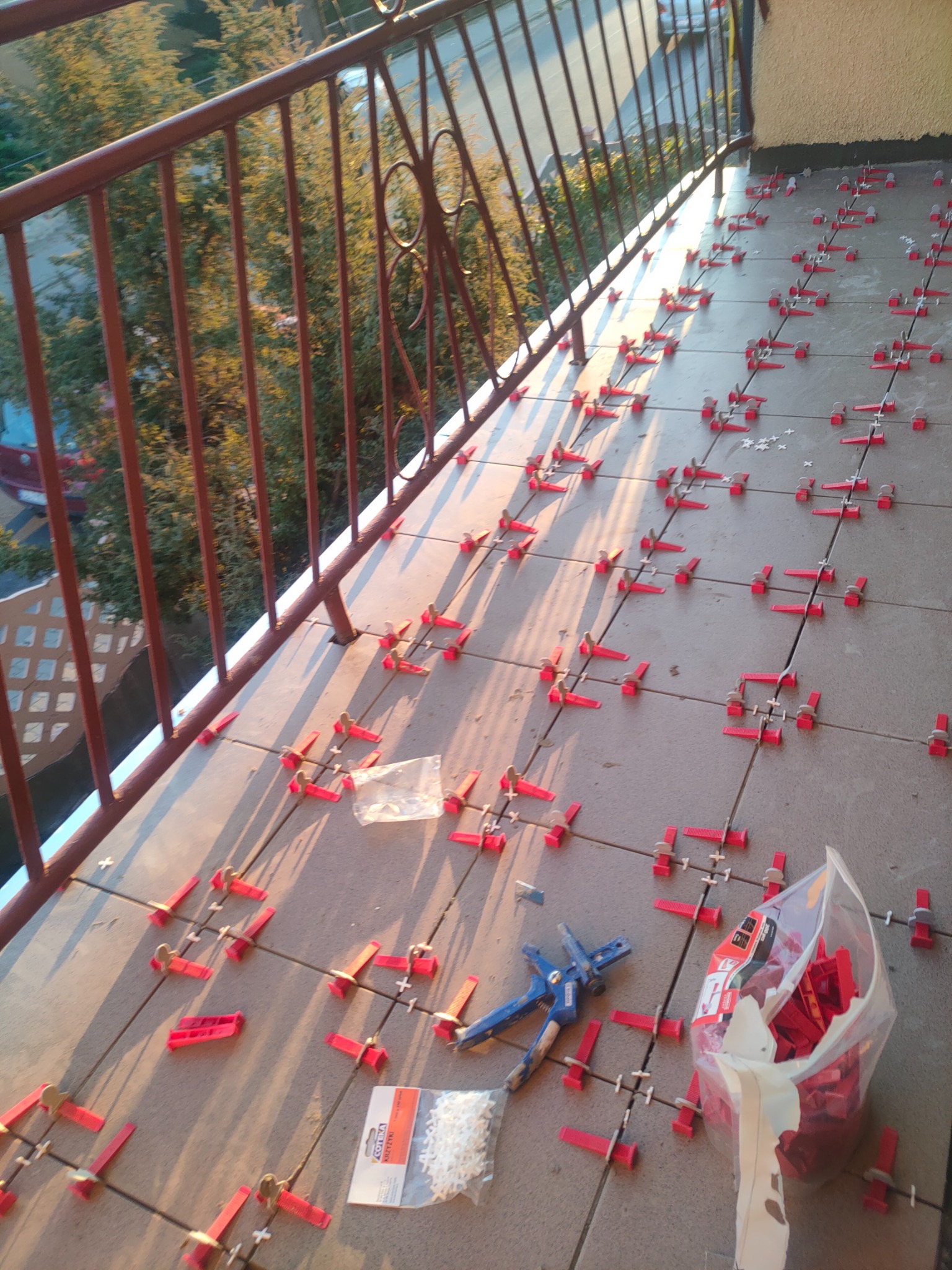 Balkon w trakcie układania płytek, widoczne narzędzia glazurnicze, system poziomowania płytek z czerwonymi klipsami, krzyżyki dystansowe, szczypce do klipsów i opakowanie z klinami.