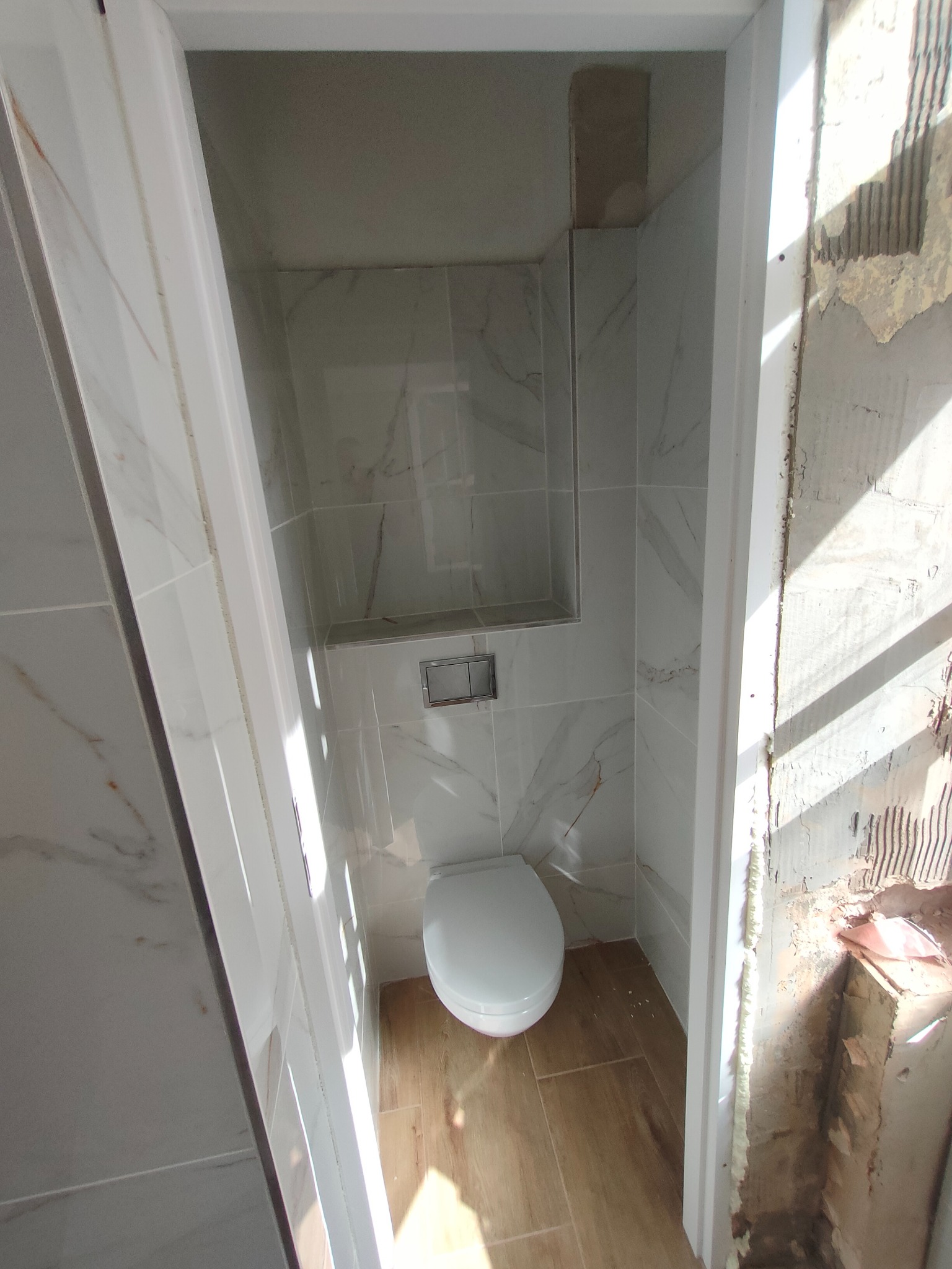 Wnętrze łazienki w trakcie remontu: biała toaleta, marmurowe płytki ścienne z wnęką, drewnopodobne płytki podłogowe, widoczne ślady po kuciu na ścianie przy futrynie drzwi.