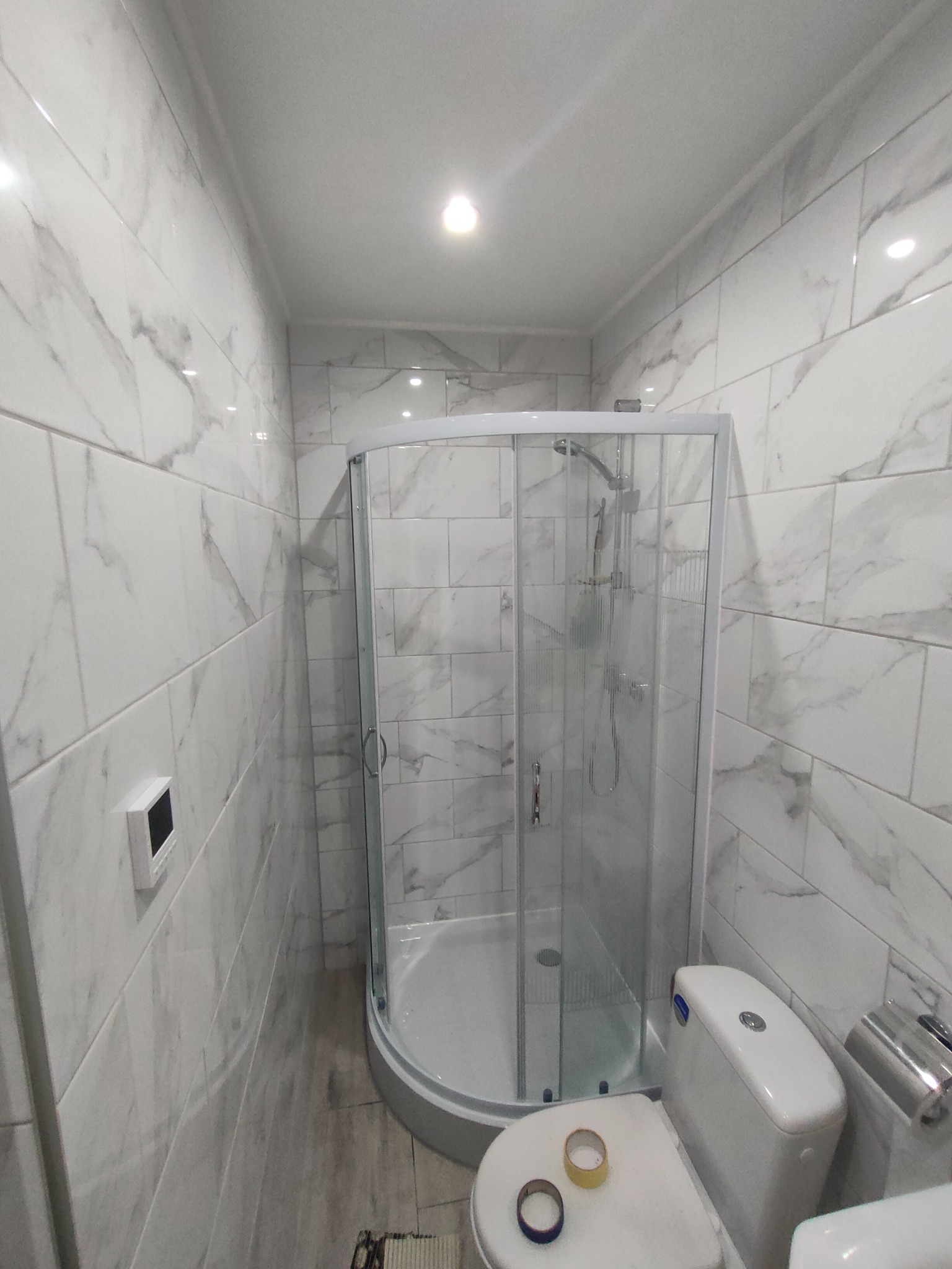 Biała łazienka po remoncie z kabiną prysznicową narożną, toaletą i płytkami imitującymi marmur na ścianach, widoczne ślady po taśmie malarskiej na desce sedesowej.