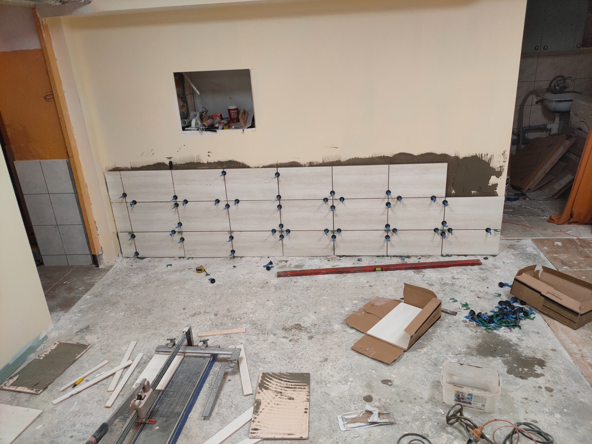 Ściana w trakcie układania płytek, widoczne niebieskie krzyżyki dystansowe, poziomica, paczka płytek i narzędzia glazurnicze na podłodze.