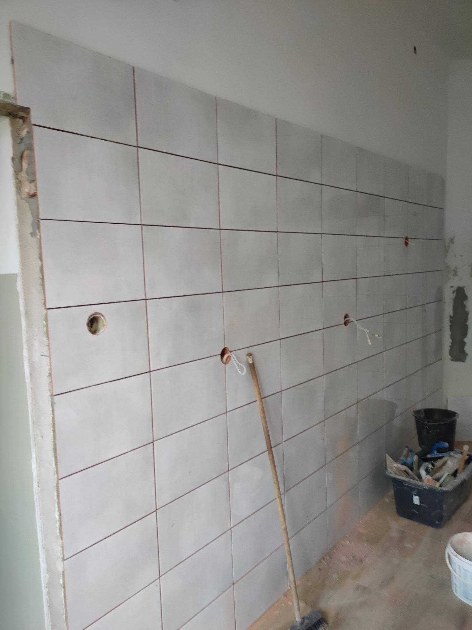 Ściana wyłożona jasnymi, kwadratowymi płytkami ceramicznymi w trakcie prac, widoczne otwory na gniazdka elektryczne z wystającymi przewodami, oraz narzędzia i wiadro na podłodze.