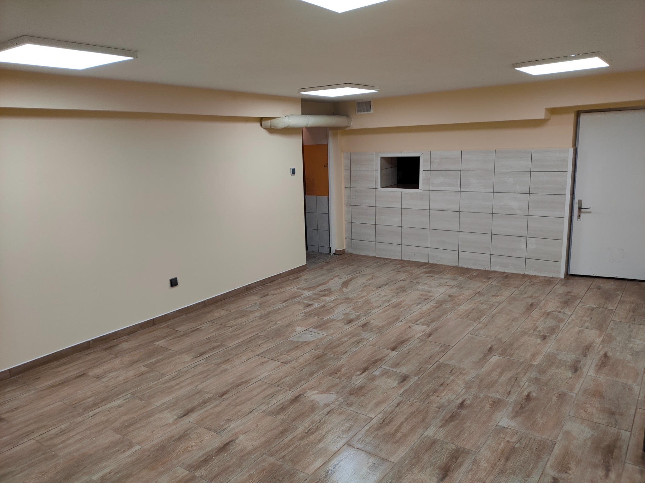 Puste pomieszczenie po remoncie z ułożoną podłogą z płytek imitujących drewno, częściowo wykafelkowaną ścianą z otworem wentylacyjnym, pomalowanymi ścianami na beżowo i białymi drzwiami.
