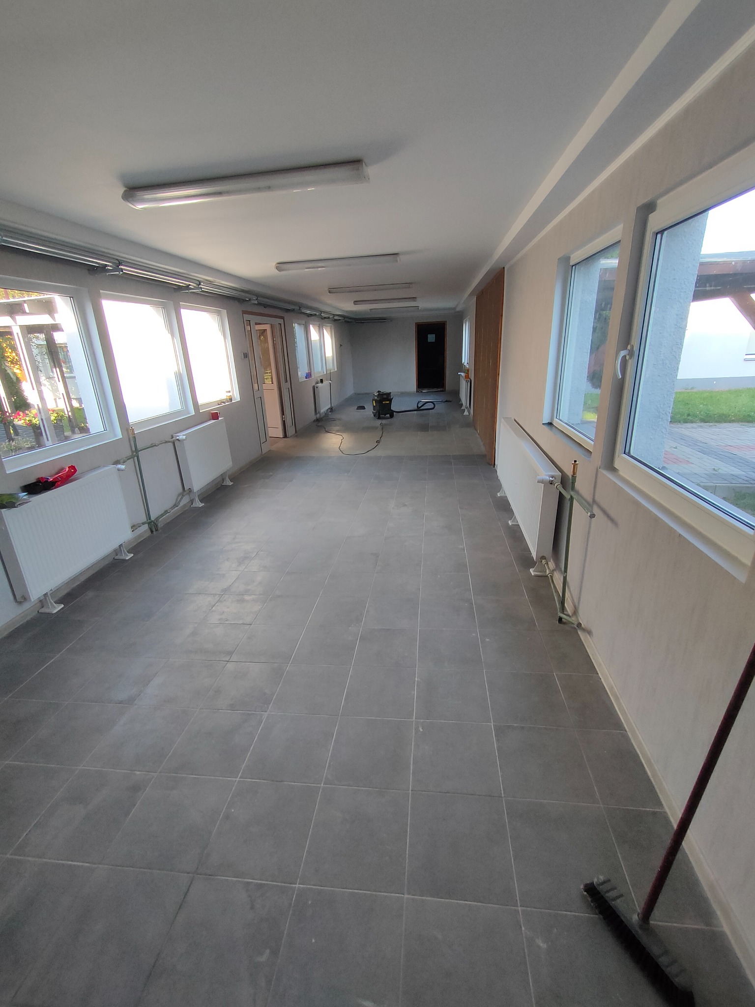 Długi korytarz biurowy po remoncie, z szarymi płytkami podłogowymi, białymi grzejnikami pod oknami i widocznym odkurzaczem przemysłowym w oddali.