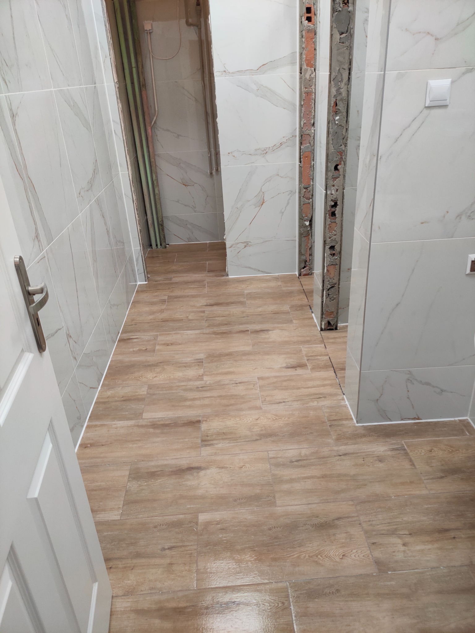 Wnętrze łazienki w trakcie remontu: położone jasne płytki imitujące marmur na ścianach i drewnopodobne na podłodze, widoczne fragmenty odsłoniętej instalacji i niedokończone obramowania drzwi.