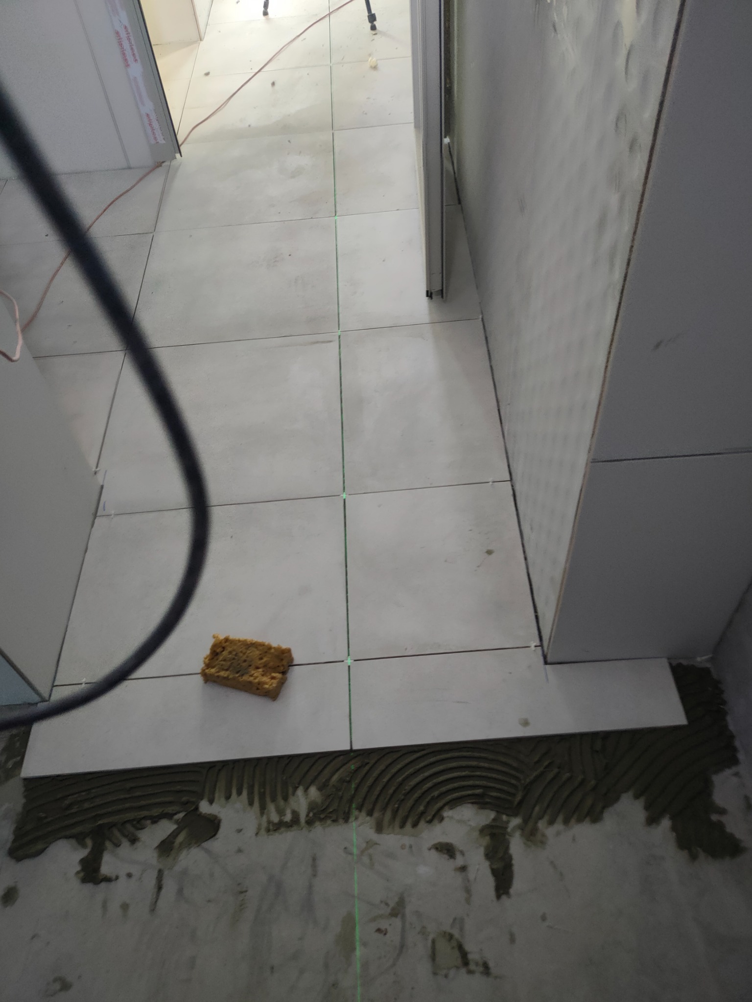 Układanie jasnych płytek podłogowych w korytarzu, widoczny pasek kleju i gąbka do czyszczenia, wyznaczony poziom lasera.