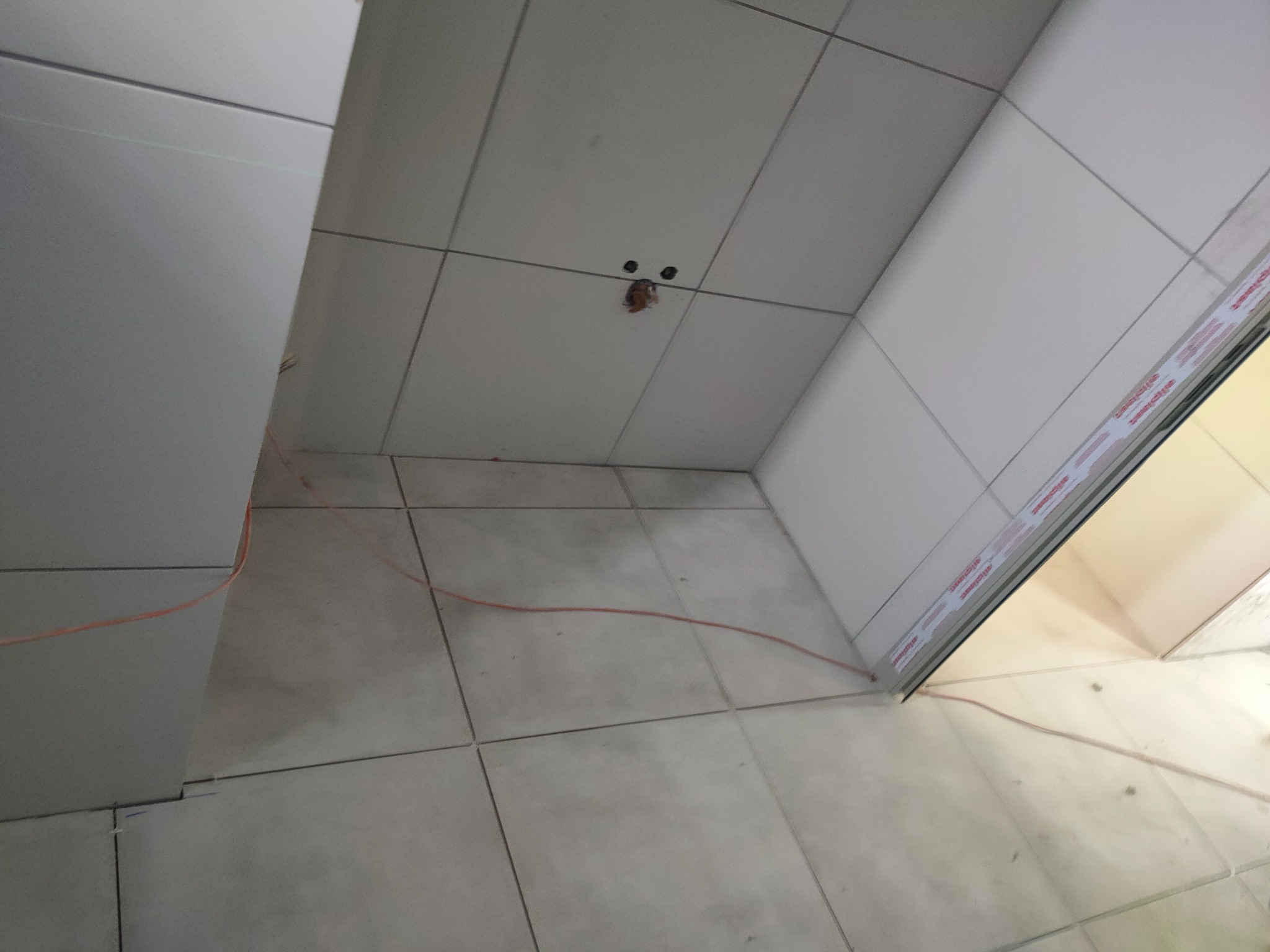 Wnętrze łazienki w trakcie remontu, widoczne białe płytki na podłodze i ścianach, z otworem w suficie i pomarańczowym przewodem elektrycznym na podłodze.