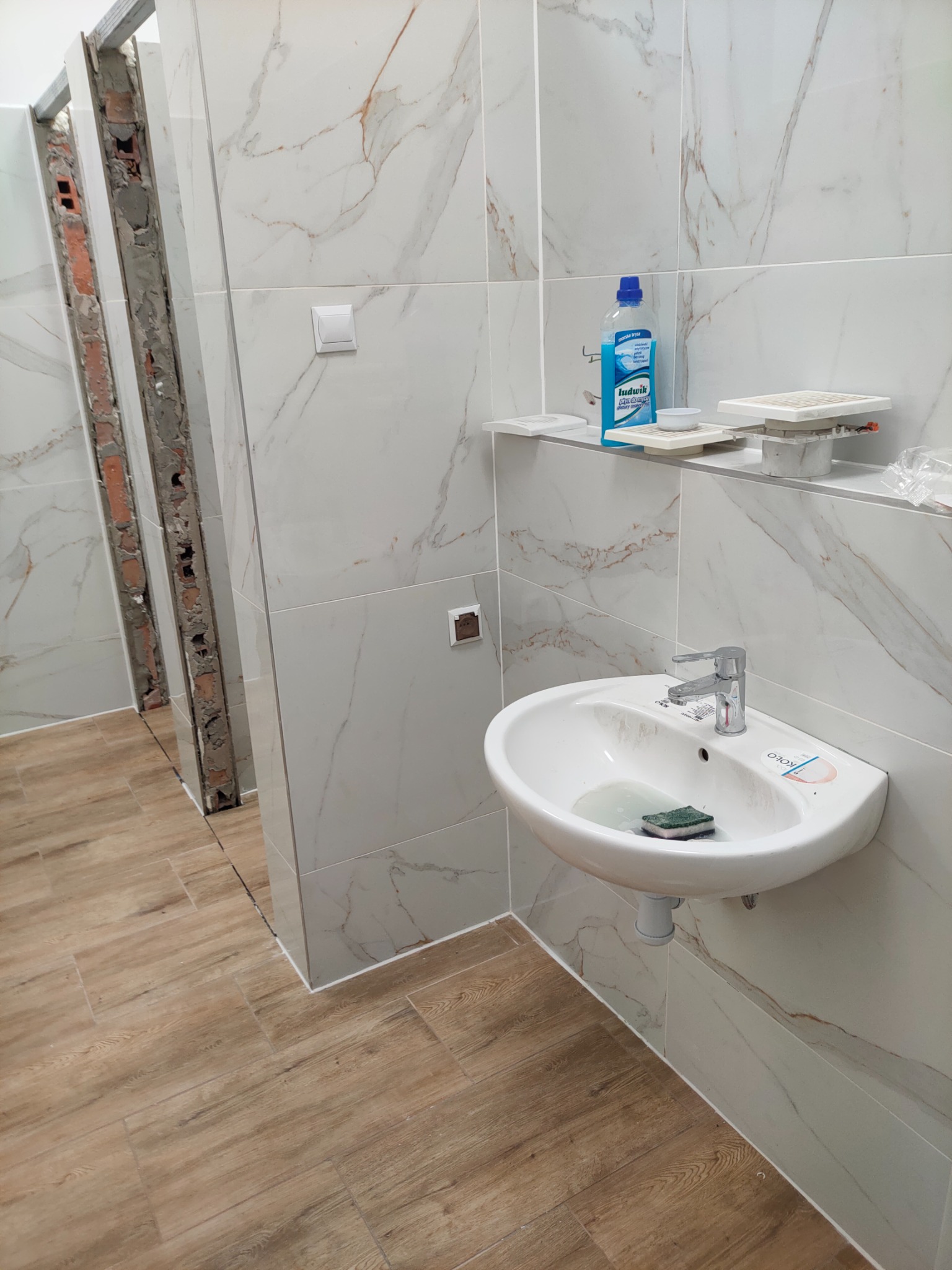 Wnętrze łazienki w trakcie prac: widoczna umywalka z baterią, drewnopodobne płytki podłogowe, marmurowe płytki ścienne, fragment niedokończonej ściany z cegłą, butelka płynu do czyszczenia na półce.