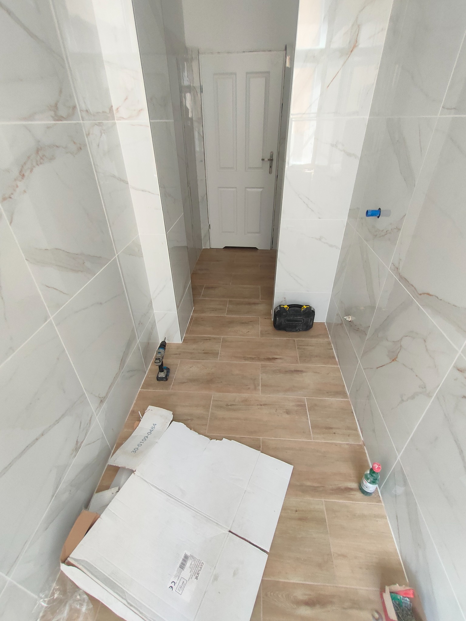 Wnętrze łazienki w trakcie wykańczania: białe, marmurkowe płytki ścienne, drewnopodobne płytki podłogowe, narzędzia pracy, kartony po płytkach i białe drzwi w tle.