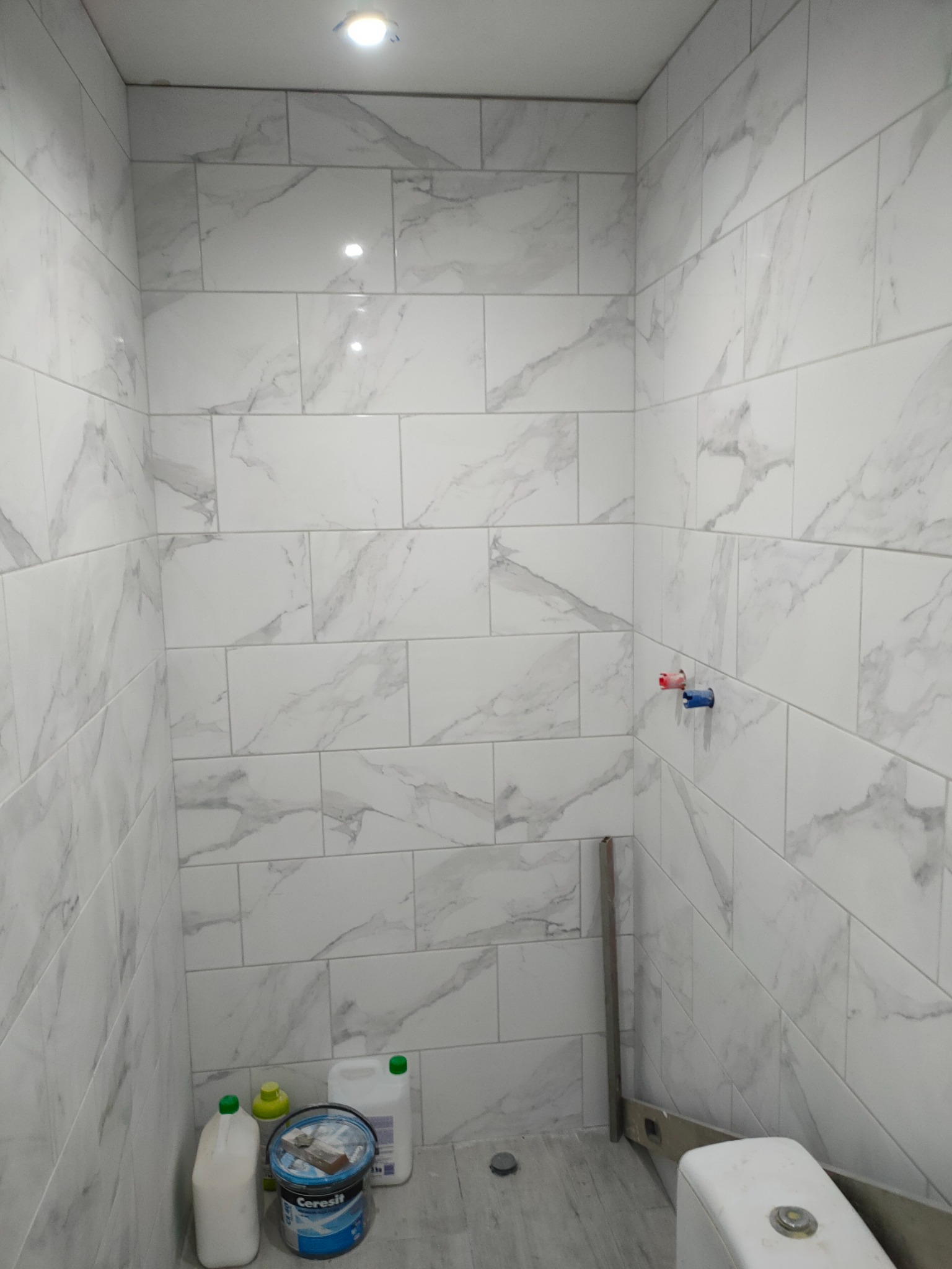 Wykończona łazienka z białymi, marmurkowymi płytkami, widoczne przyłącza wodne, poziomica oparta o ścianę i częściowo widoczna toaleta, na podłodze wiadro z klejem do płytek marki Ceresit.
