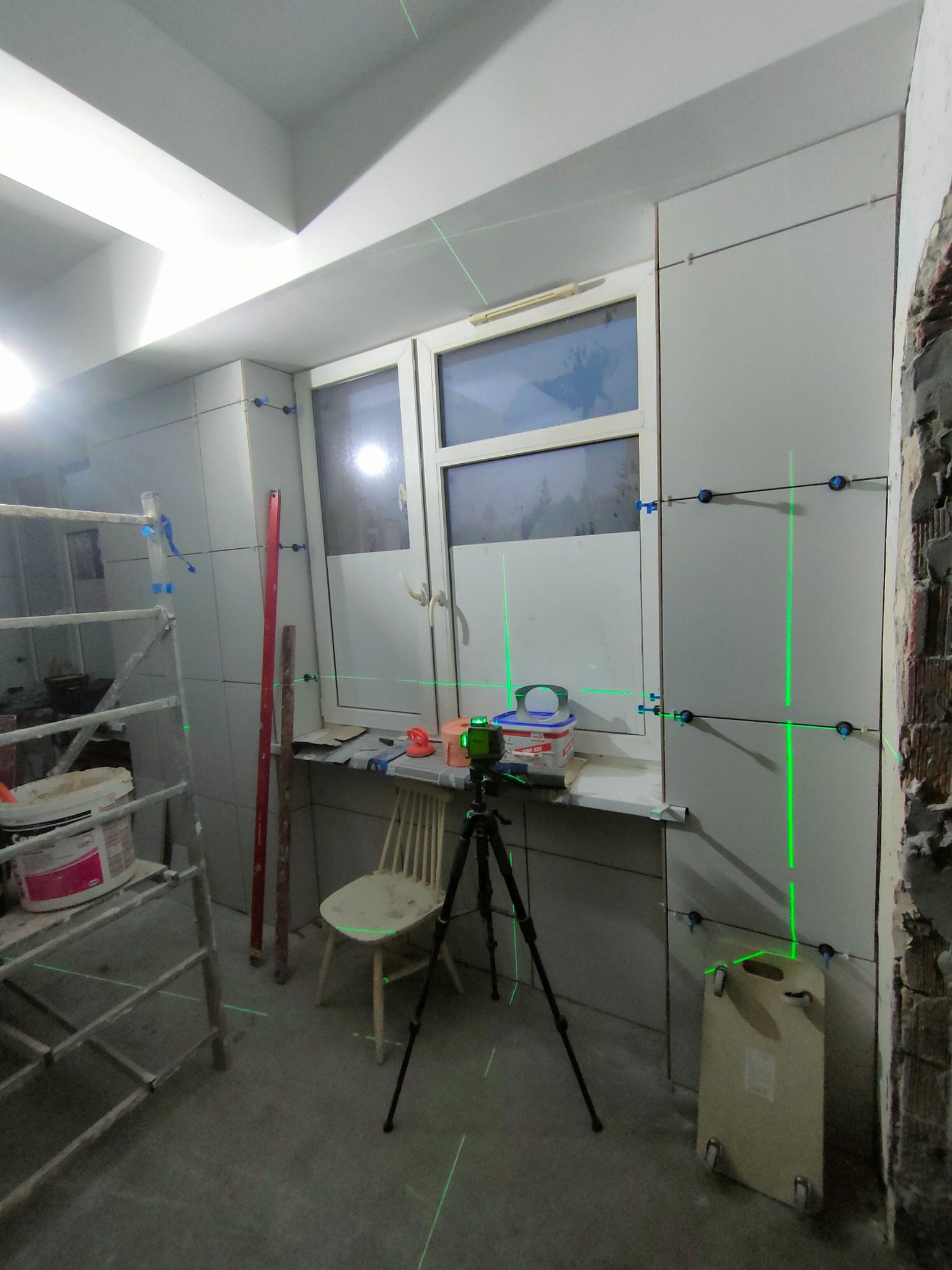 Ściana w trakcie układania płytek, widoczne poziomice laserowe, krzyżyki dystansowe, drabina i niedokończone fragmenty ściany, w tle okno.