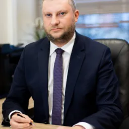 Mężczyzna w garniturze siedzi przy biurku, trzyma długopis nad dokumentem z tabelą, patrząc wprost w obiektyw.